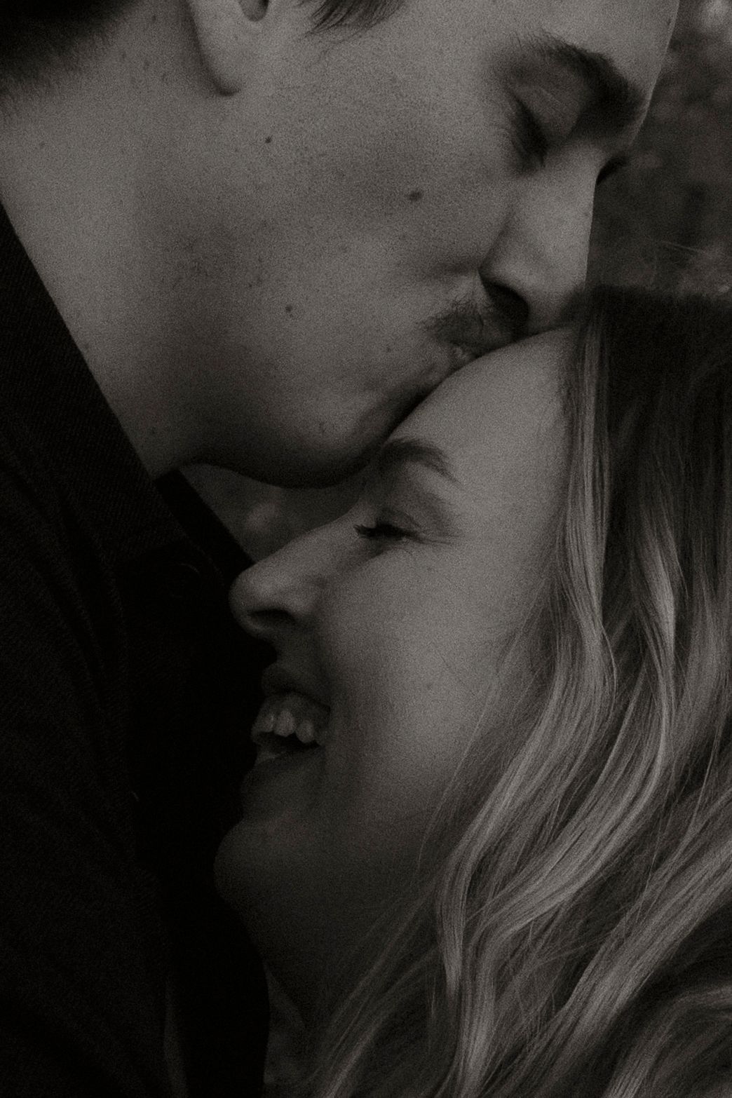 Black and white photo of a couple sharing a tender kiss, woman smiling warmly as man kisses her forehead.