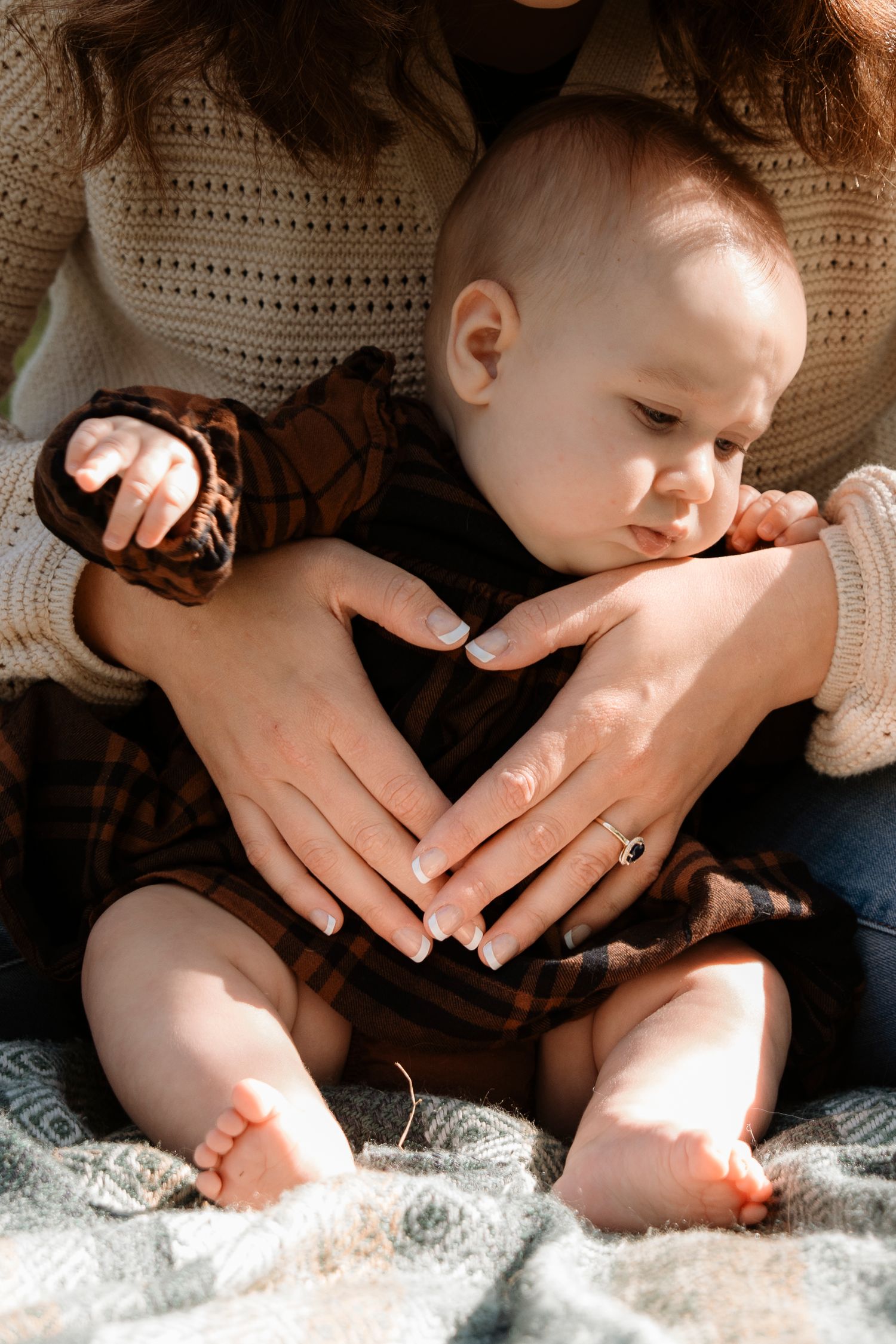 A mother gently holds her baby in her lap outdoors, with the infant wearing a plaid outfit and tiny bare feet visible.