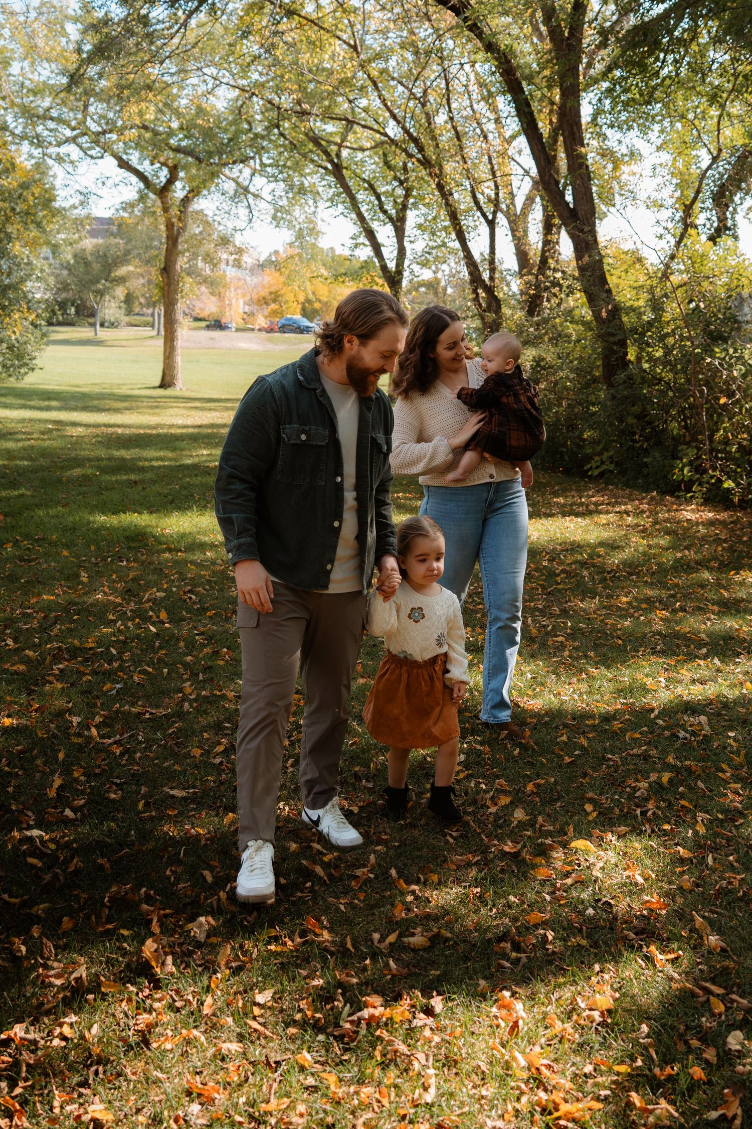 Family of four enjoying autumn park, parents with toddler and baby among fallen leaves and trees.
