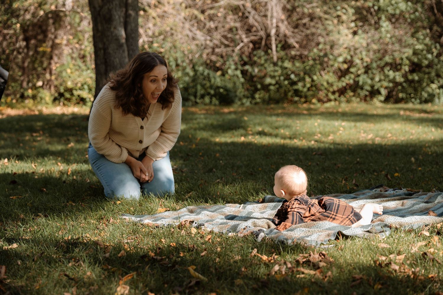 A mother kneels on grass watching her baby lying on a blanket outdoors in a sunlit autumn park.