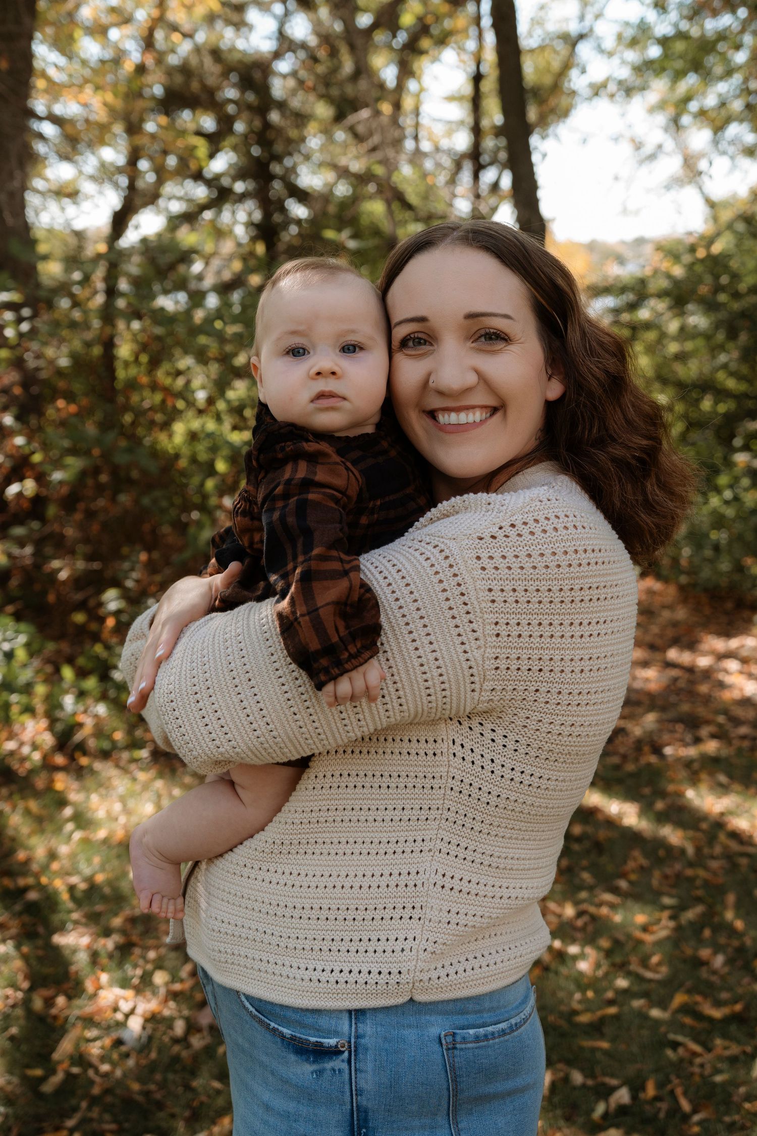 Smiling woman in cream knit sweater holds baby in plaid shirt outdoors in autumn forest setting.