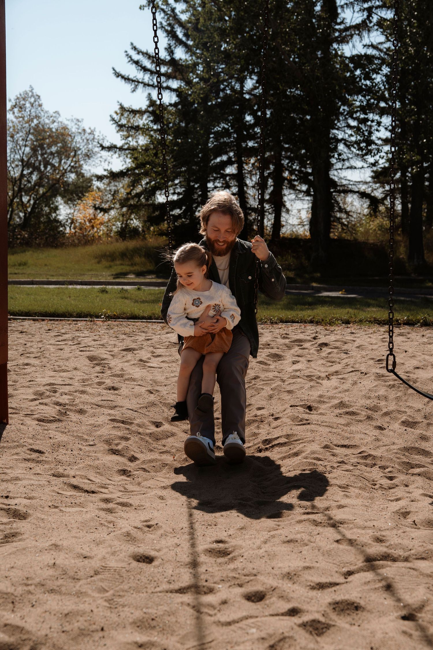 Father holding toddler on playground swing set in sandy park surrounded by tall trees on sunny day.