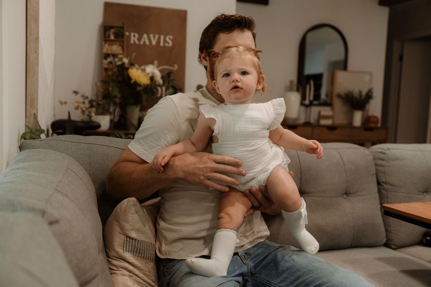 A father holds his baby girl on a cozy gray sofa in a warm living room setting.