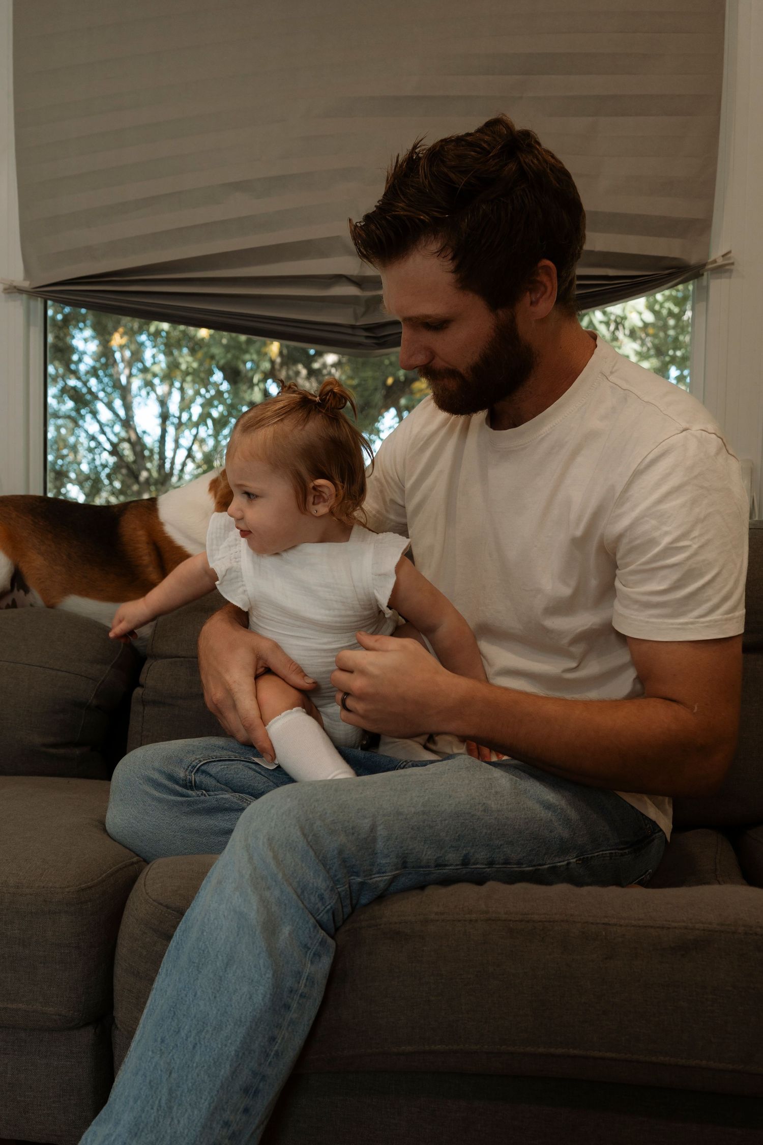 Bearded father in white t-shirt and jeans sitting on gray sofa holding baby girl in white onesie at home.