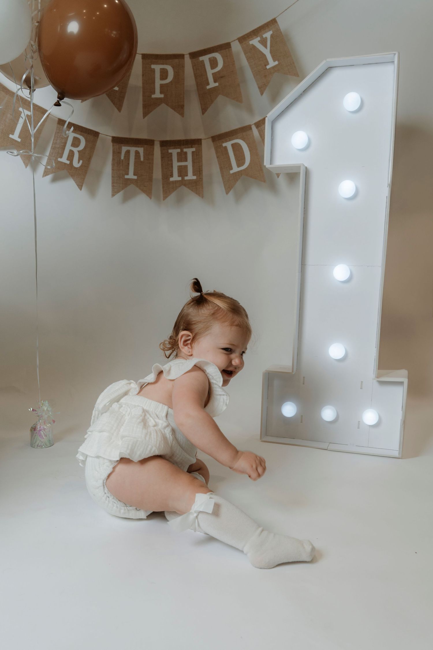 Baby girl in white romper celebrates first birthday with lit number 1, balloon, and Happy Birthday banner.