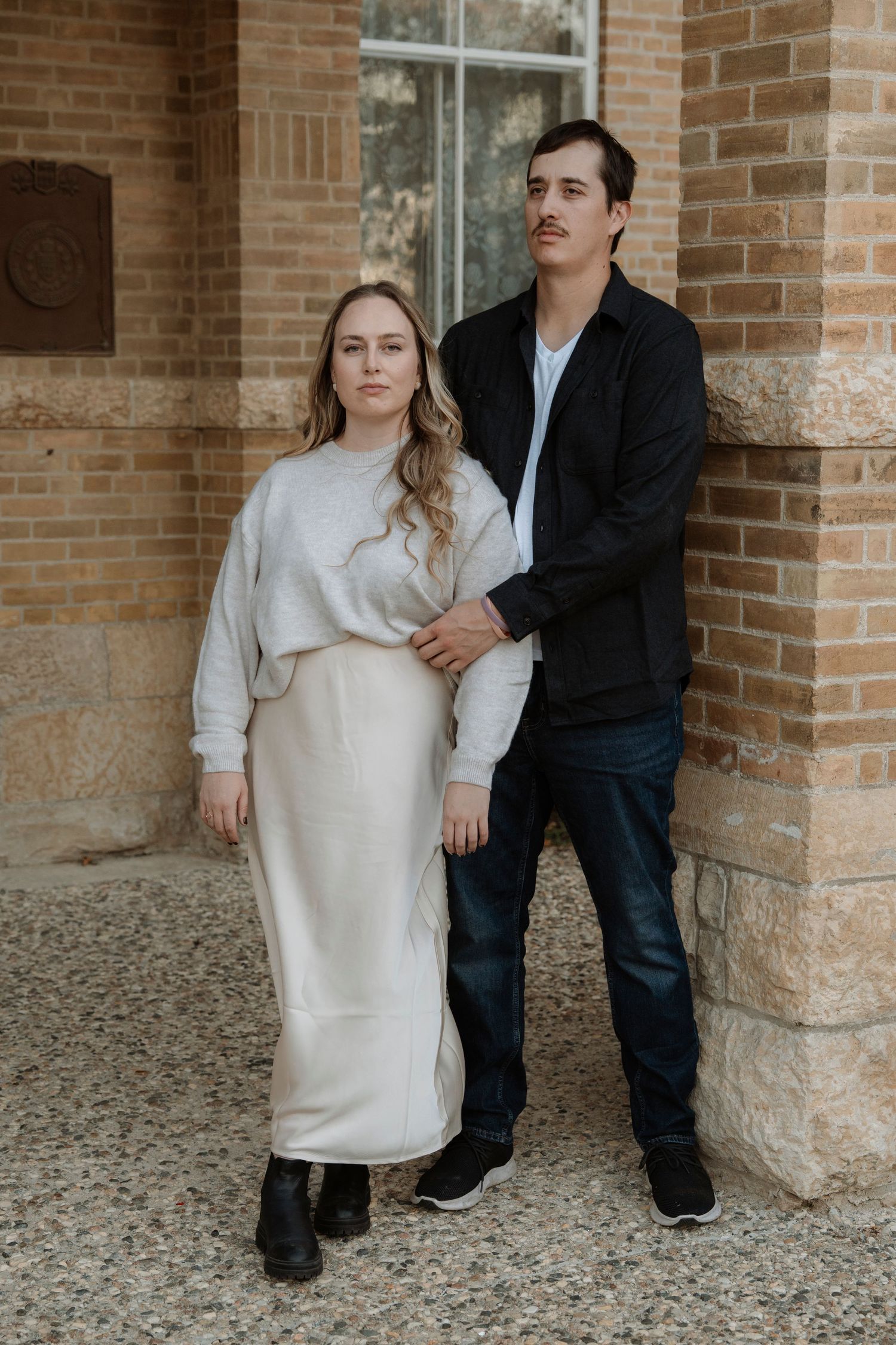 Couple posing together in front of a brick building, woman in cream skirt and man in black jacket.