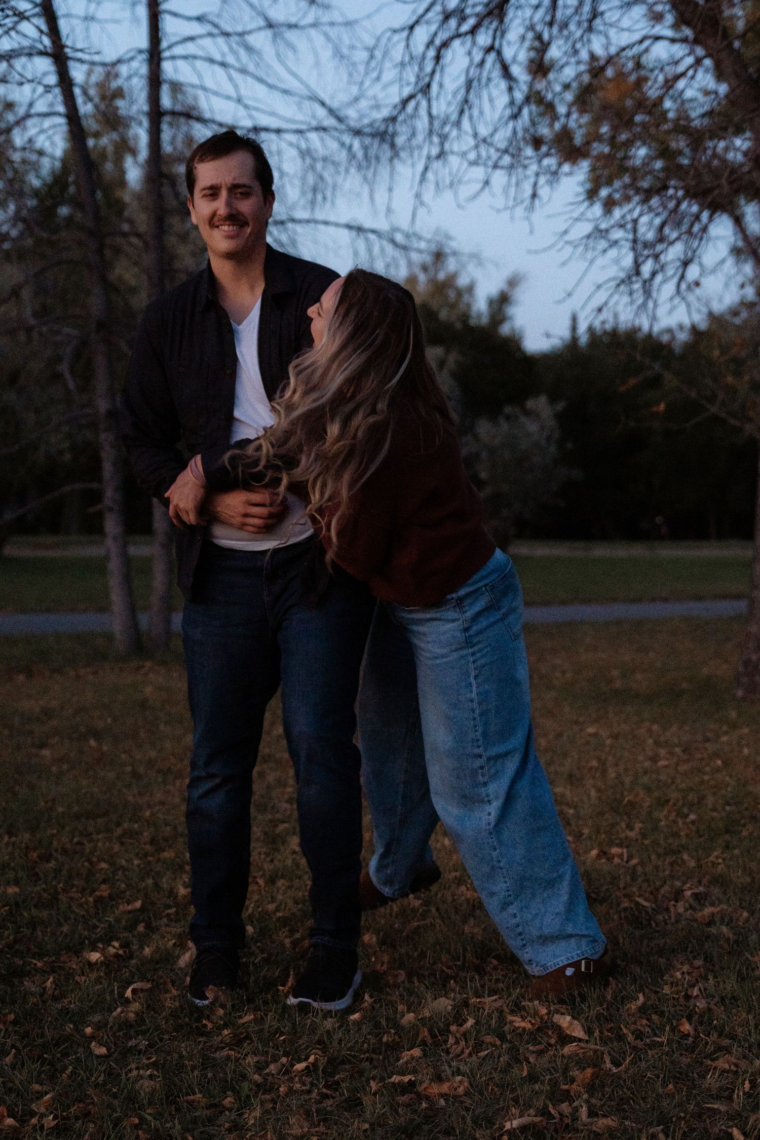 Couple laughing and embracing outdoors in a park during dusk, wearing casual fall outfits.