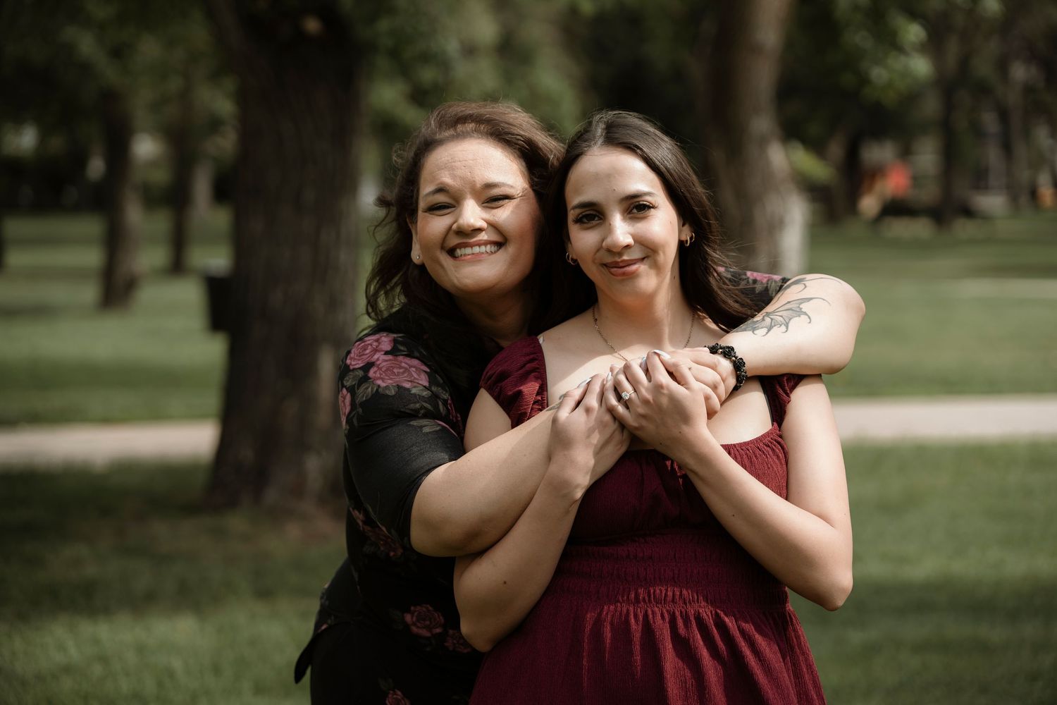 Two women in burgundy outfits embrace warmly in a lush green park, smiling joyfully at the camera.
