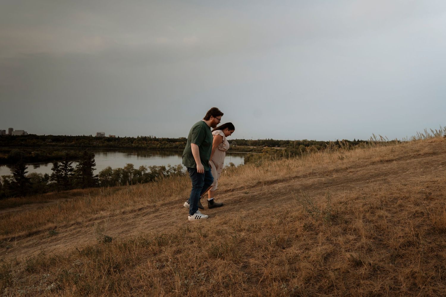 Couple walking hand in hand through golden field near a lake under overcast skies.