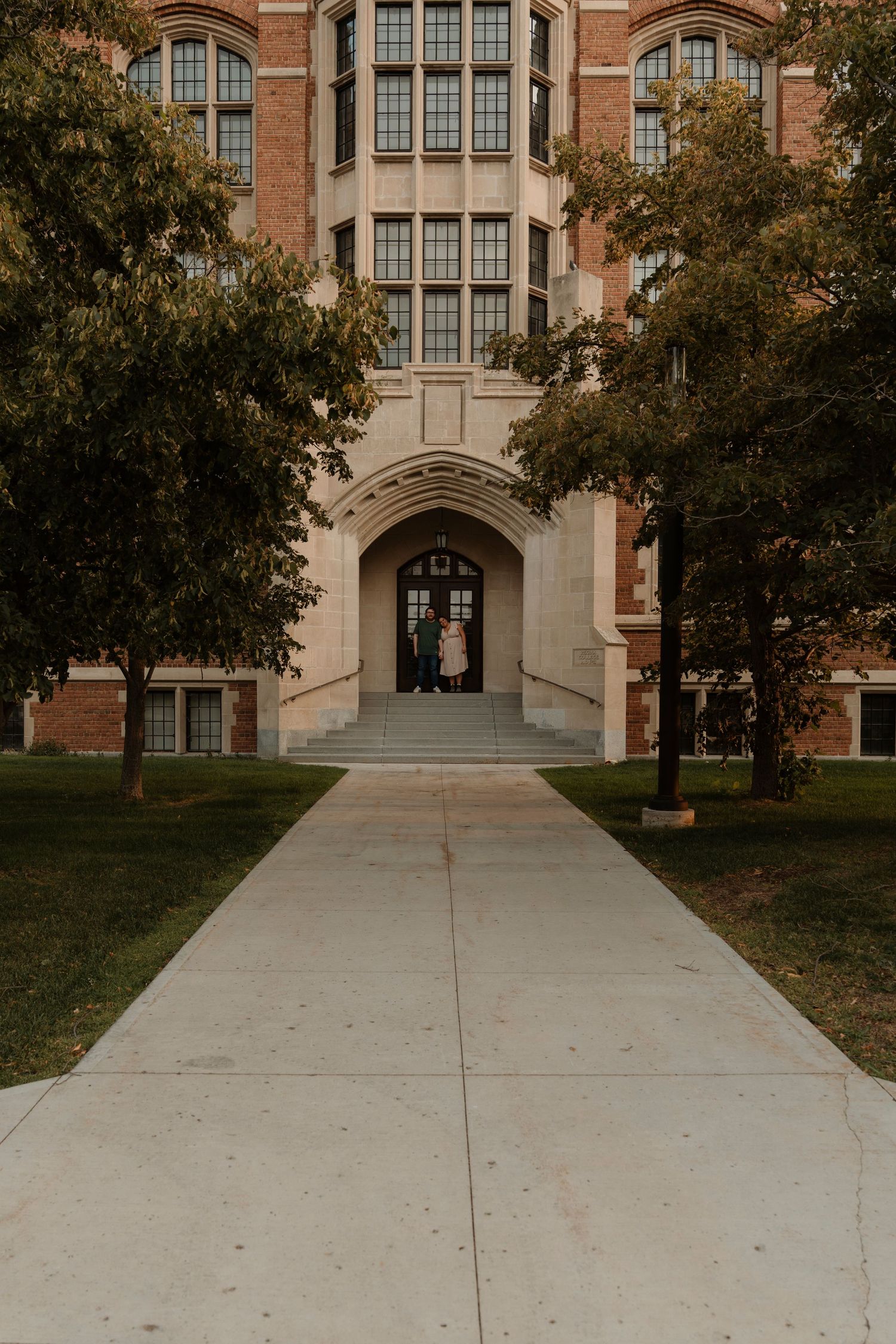 Brick university building with arched entrance, tree-lined walkway, and couple standing at the steps on a cloudy day.