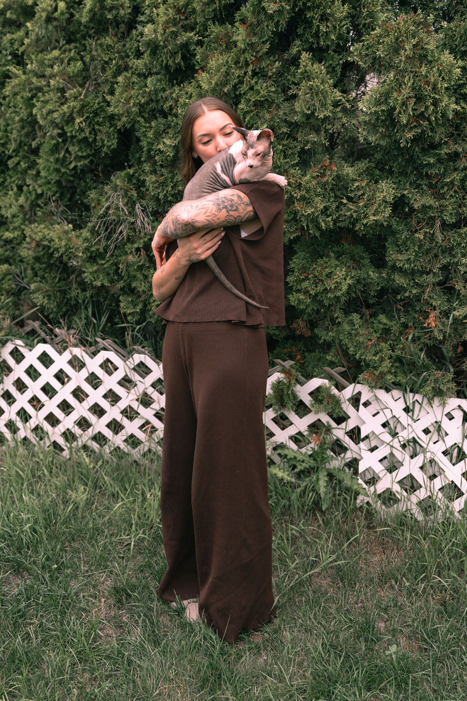 Woman in brown outfit holding a Sphynx cat outdoors near green hedges and white lattice fence.