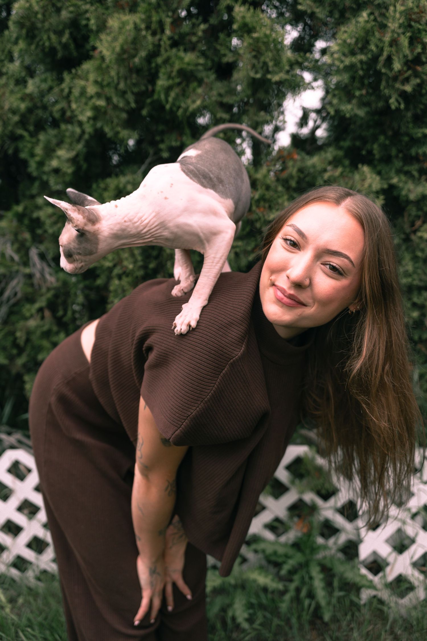 A smiling young woman in a brown dress balances a hairless Sphynx cat on her shoulder outdoors near green trees.