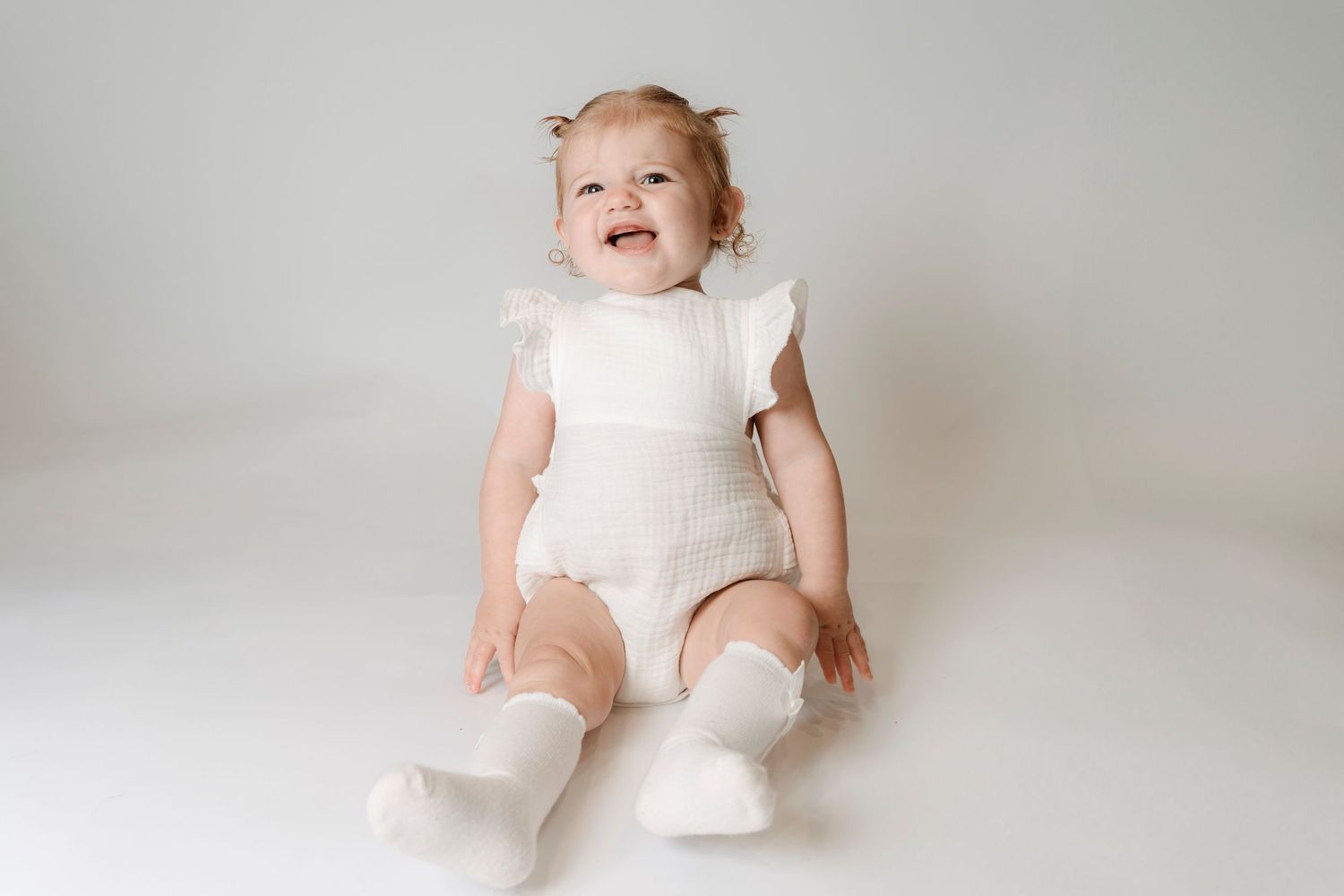 Smiling baby girl in white ruffle romper and socks sitting on white background.