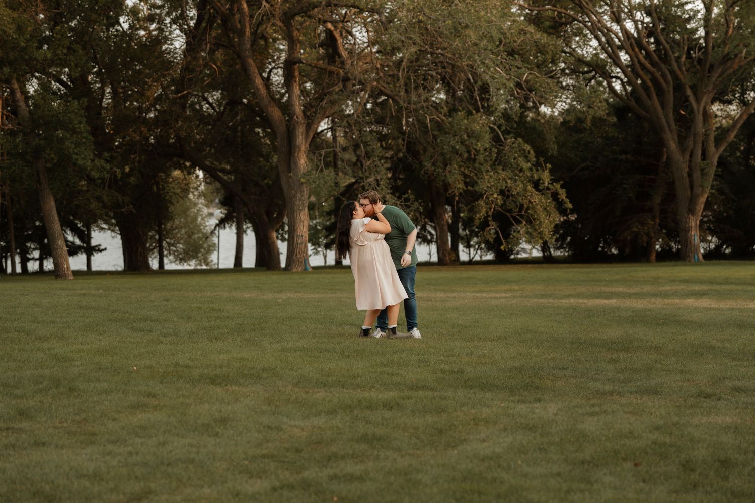 Couple embracing and kissing in a lush green park surrounded by tall trees during an outdoor engagement session.