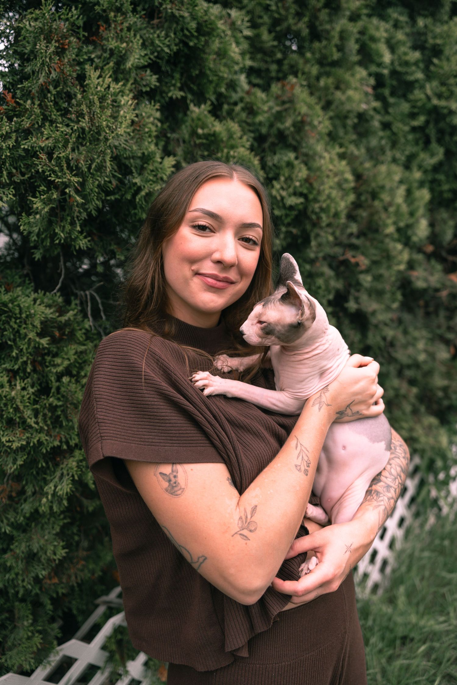Young woman with tattoos smiling outdoors, holding a hairless Sphynx cat against evergreen trees.