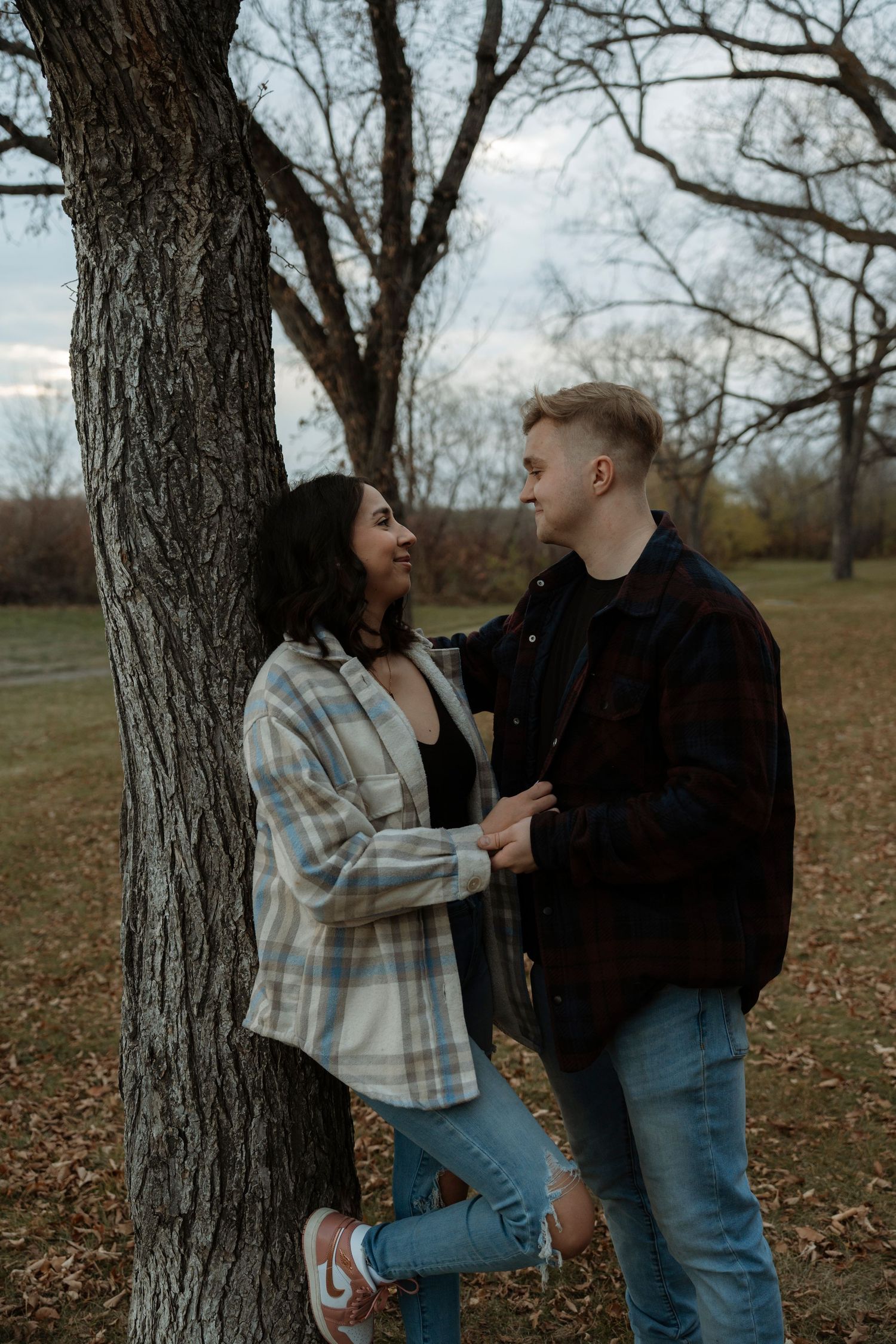 Couple sharing a romantic moment by a tree in an autumn park, wearing casual flannel shirts and jeans.