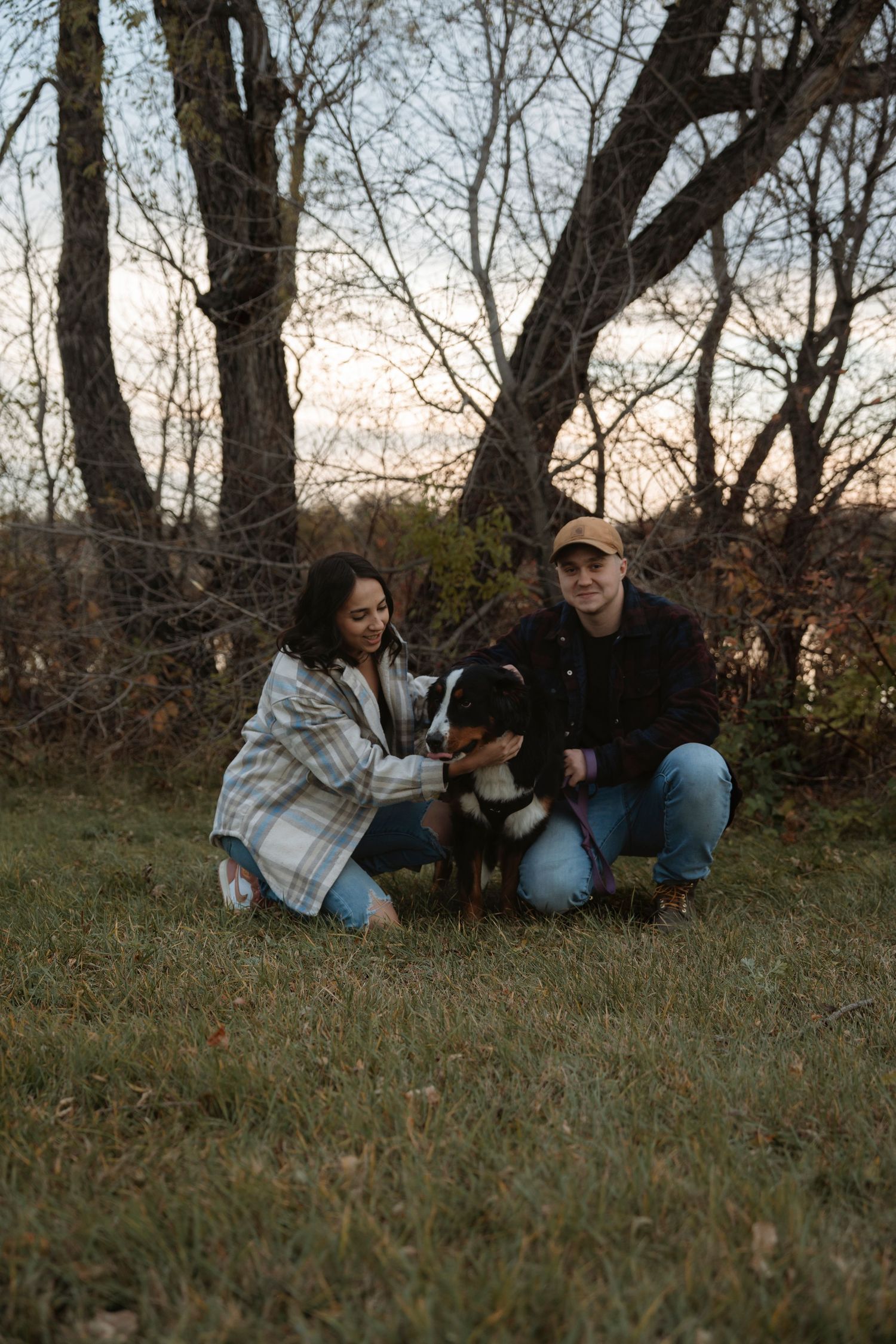 A couple crouches in a grassy field with their Bernese Mountain Dog, surrounded by bare autumn trees at dusk.