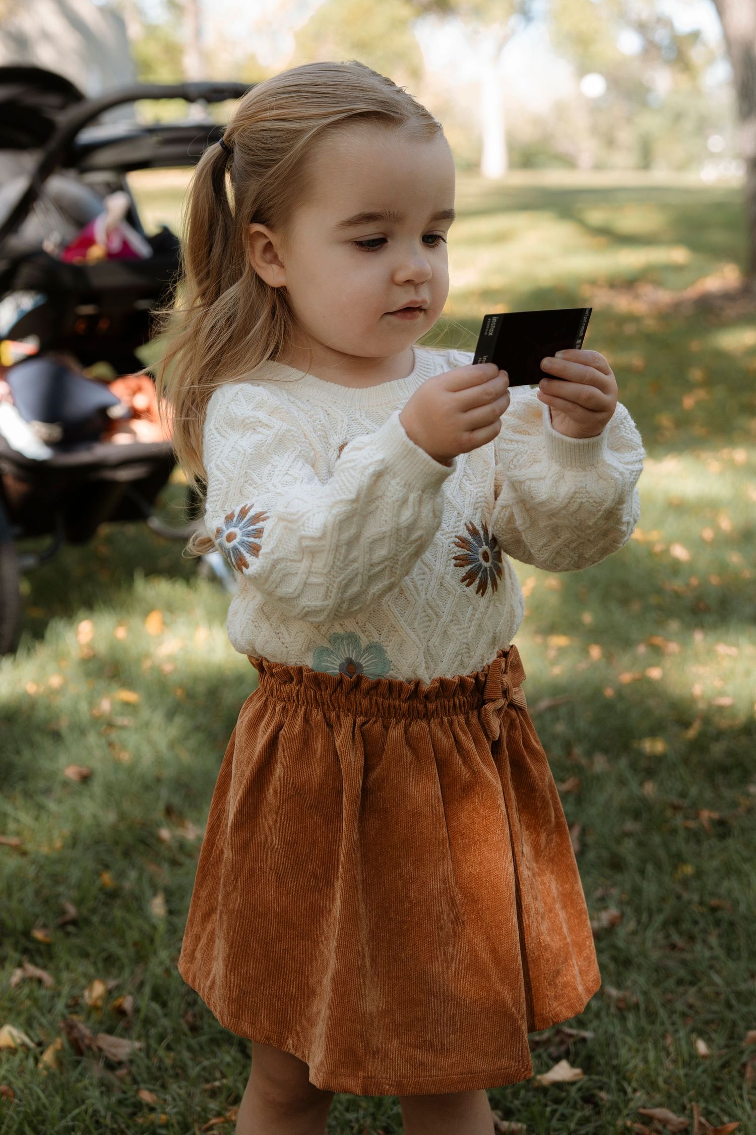 Toddler girl in white floral sweater and rust corduroy skirt holds a card outdoors in a green park.