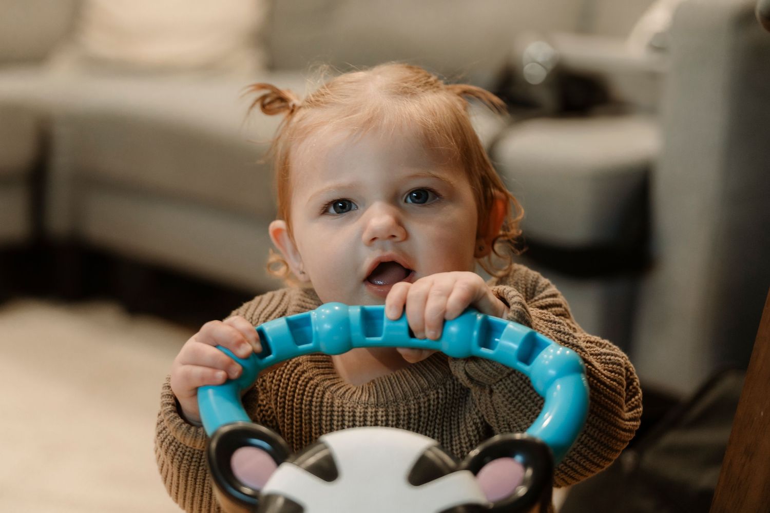 Cute toddler with pigtails holding a toy steering wheel in a cozy living room.