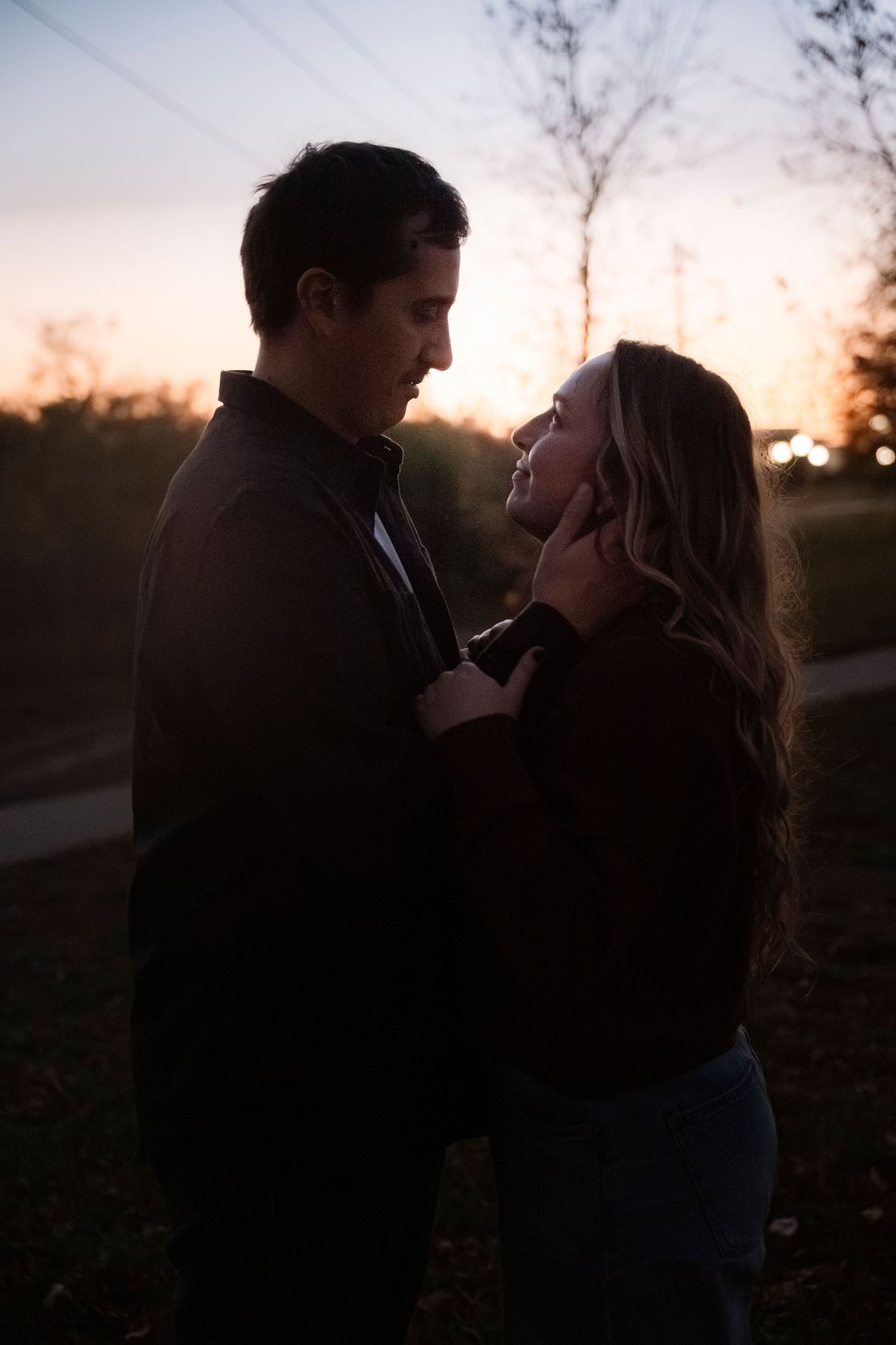 Romantic couple embracing at sunset in a park, silhouetted against golden sky, woman smiling up at man tenderly.