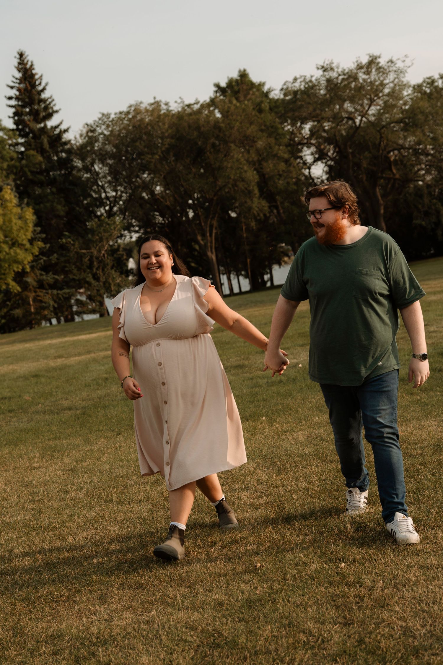 A happy couple holds hands and walks through a green park, smiling at each other on a warm summer evening.