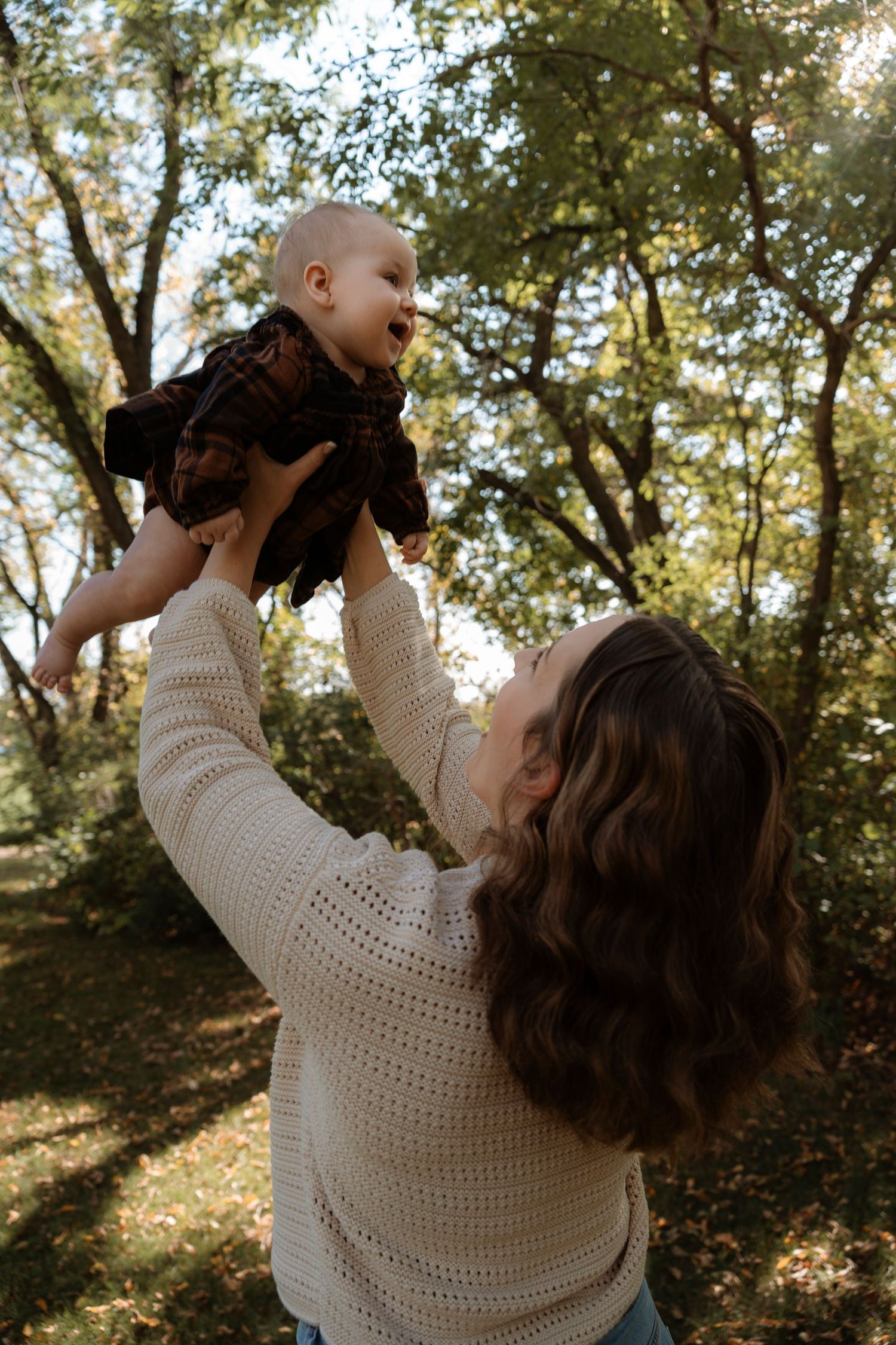 A mother lifts her smiling baby overhead in a sunlit park, both wearing cozy autumn outfits among green trees.