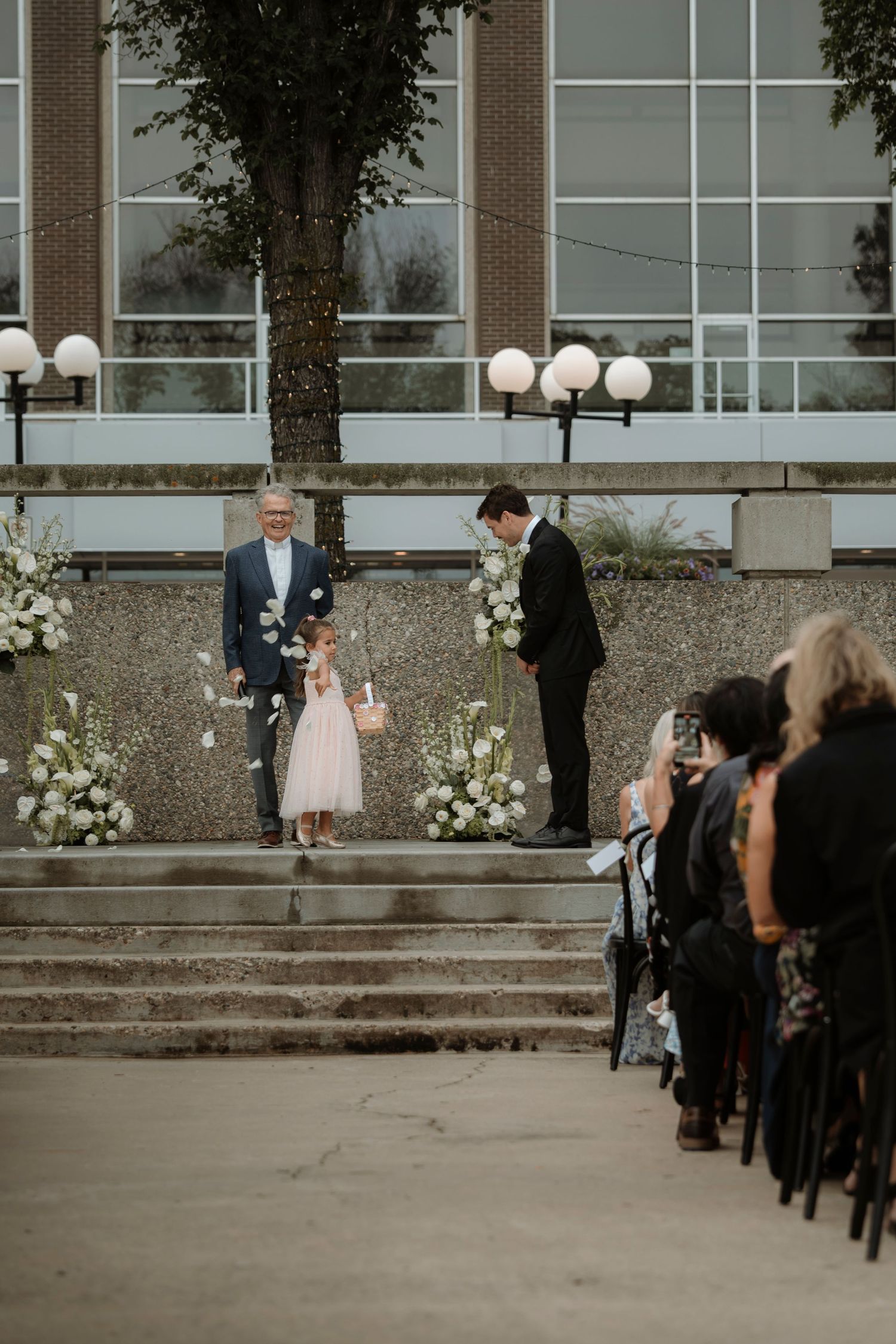 Flower girl walks down outdoor wedding aisle with white floral arrangements as groom waits at the altar.