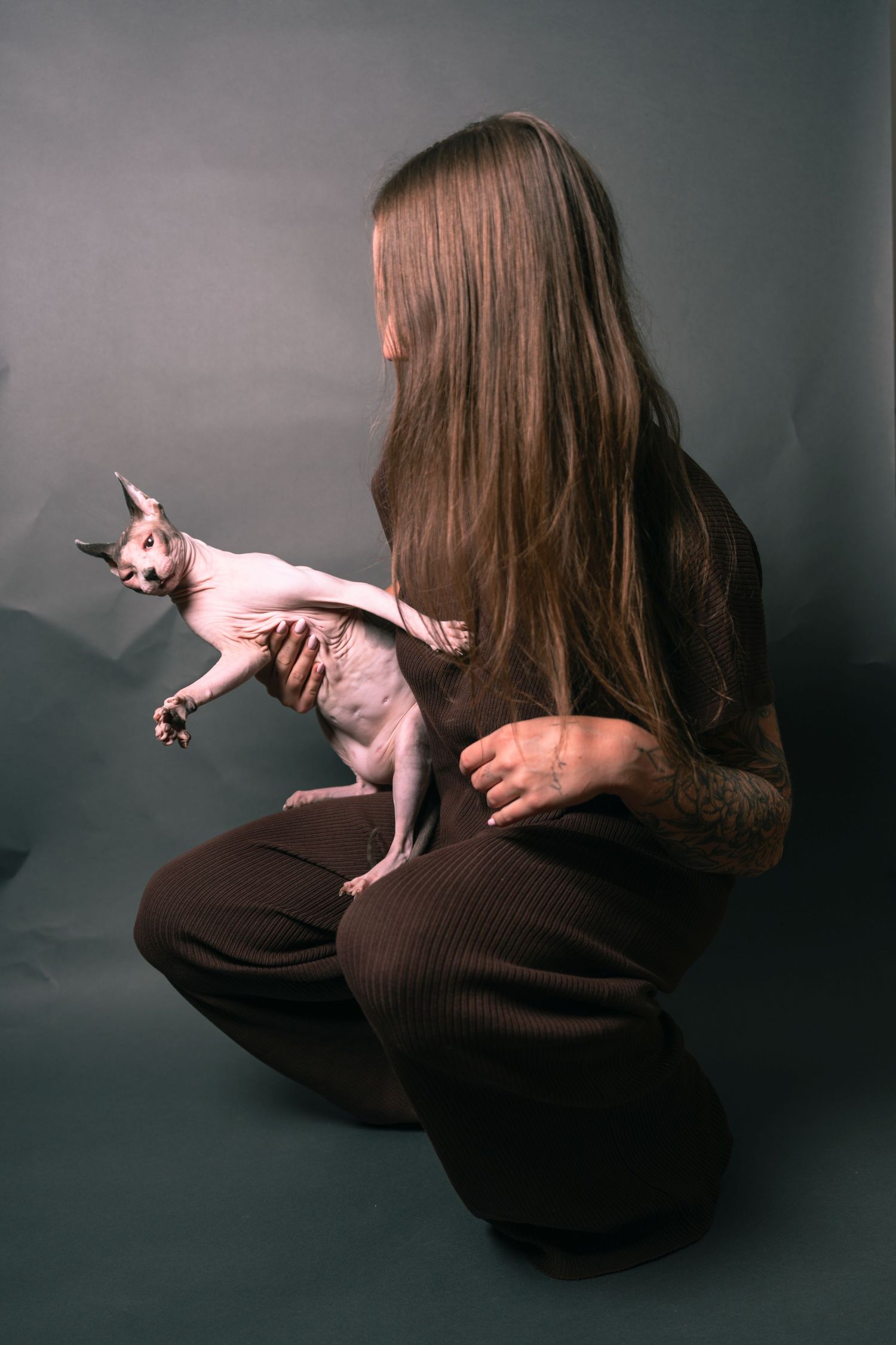 A woman with long hair sits cross-legged holding a Sphynx cat against a dark gray background.