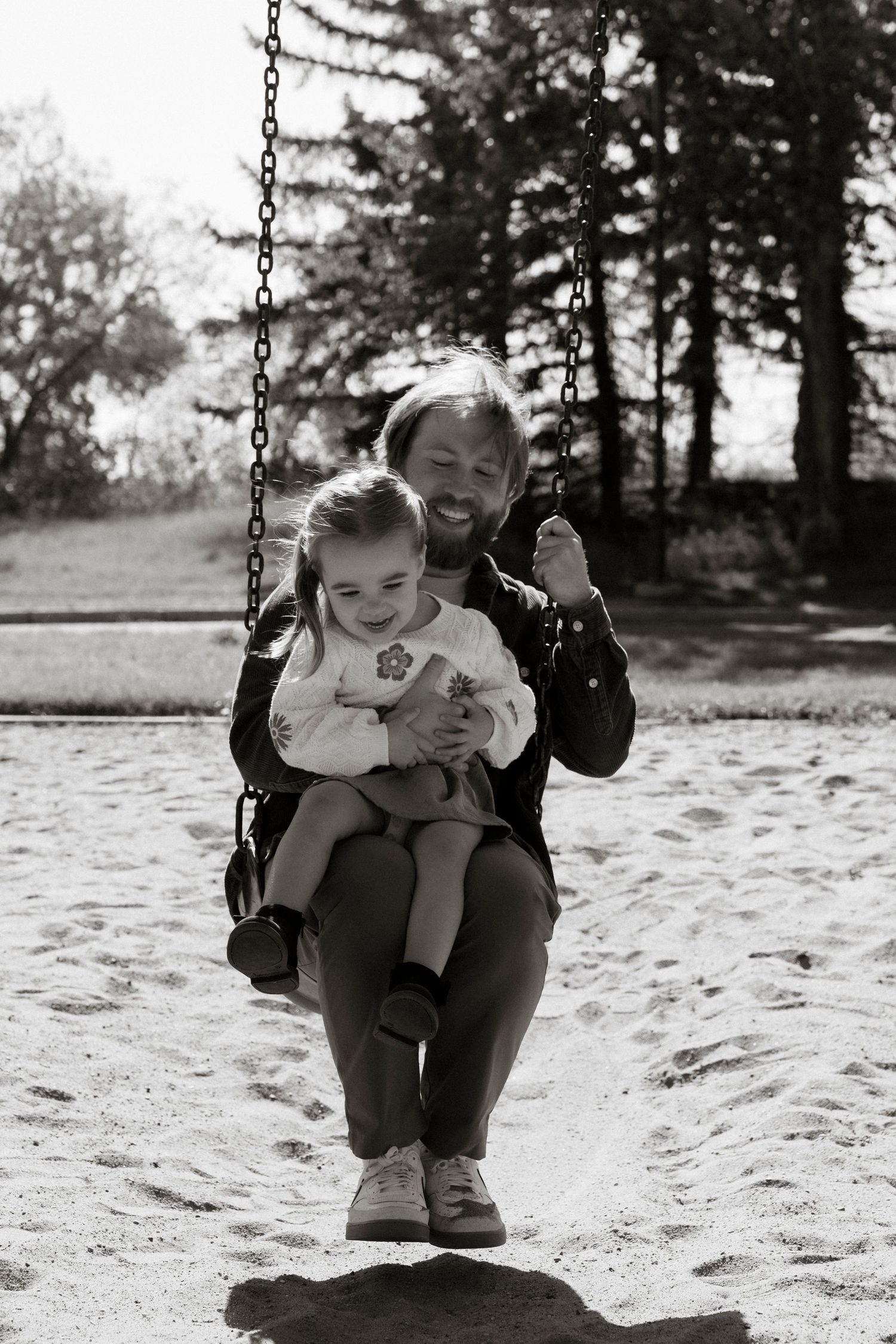 Black and white photo of a man and young girl smiling together on a playground swing, surrounded by trees.