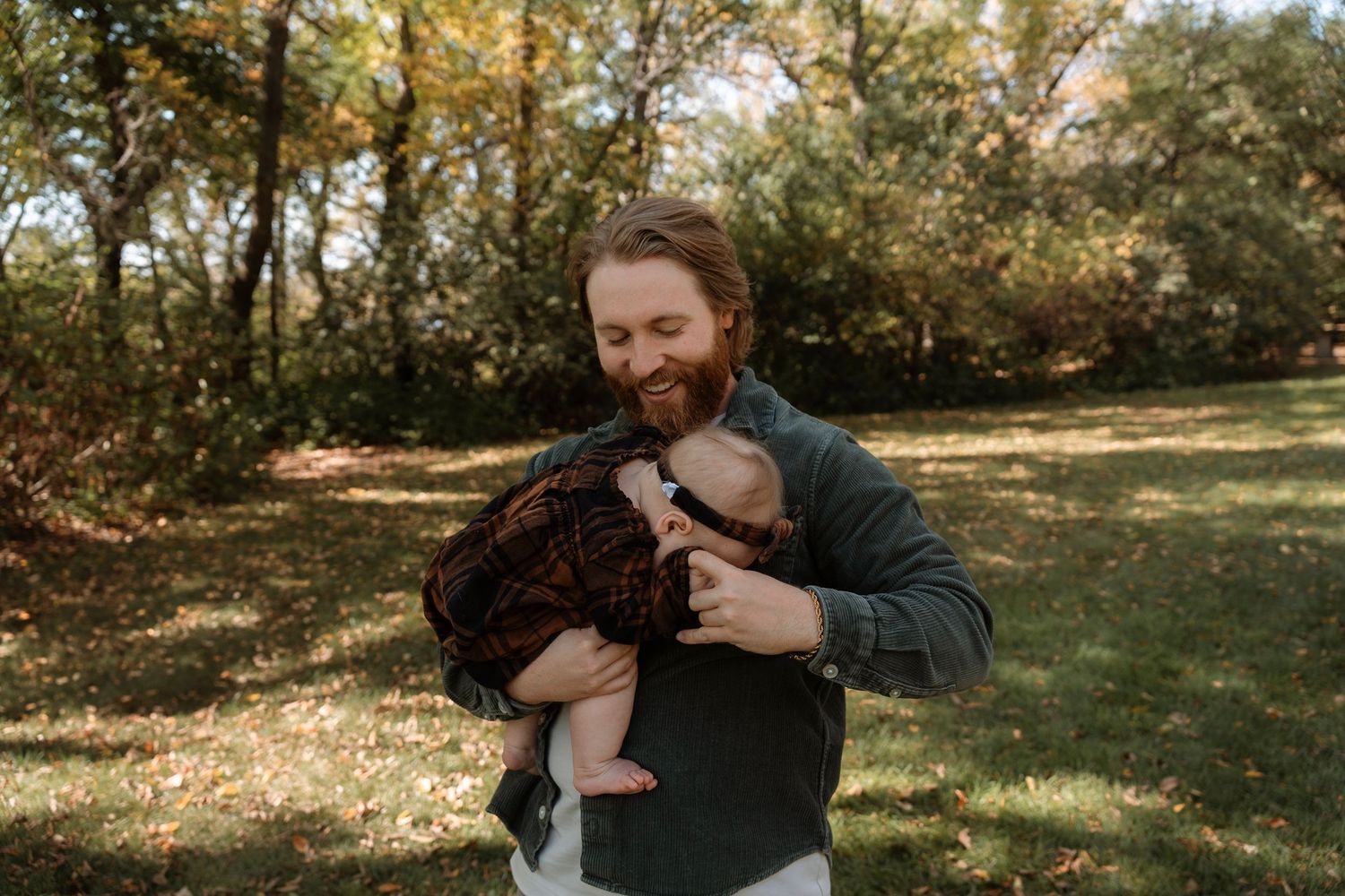 A smiling bearded man holds a baby in a dark outfit outdoors in a lush green park setting.
