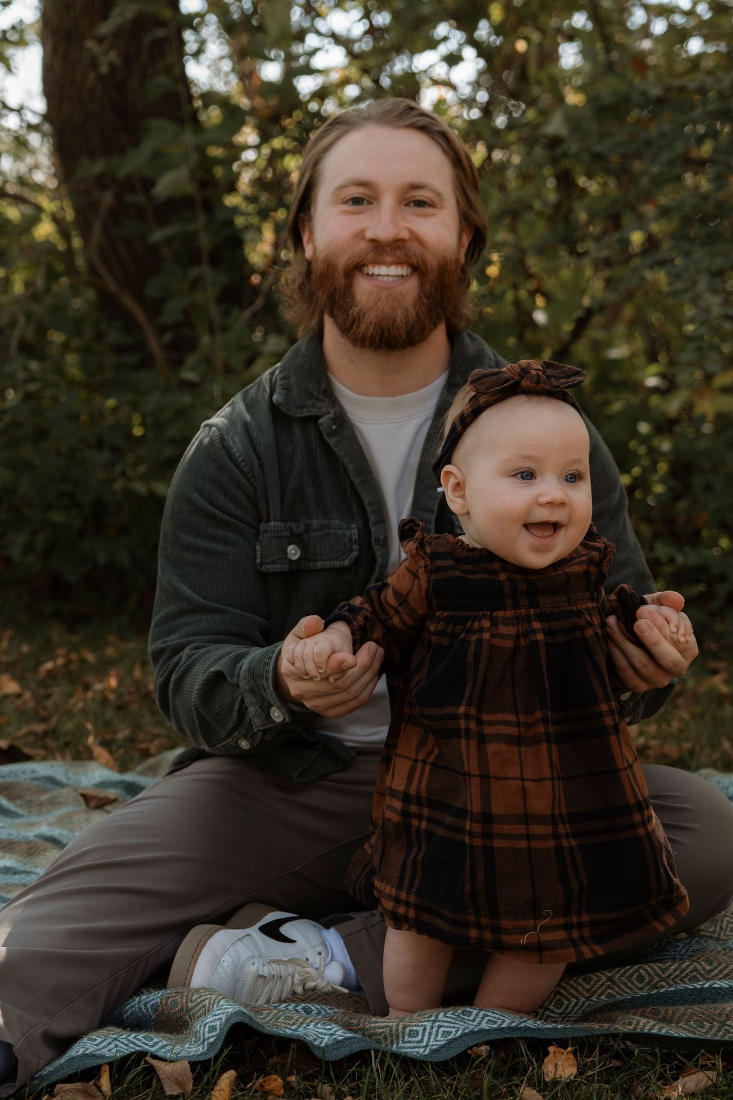 Smiling bearded man holding a happy baby in a brown plaid dress outdoors on a blanket among autumn leaves.