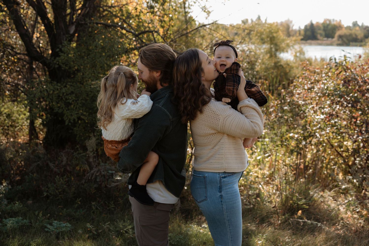A family of four enjoys a warm autumn day outdoors, with parents kissing while each holding a young child.