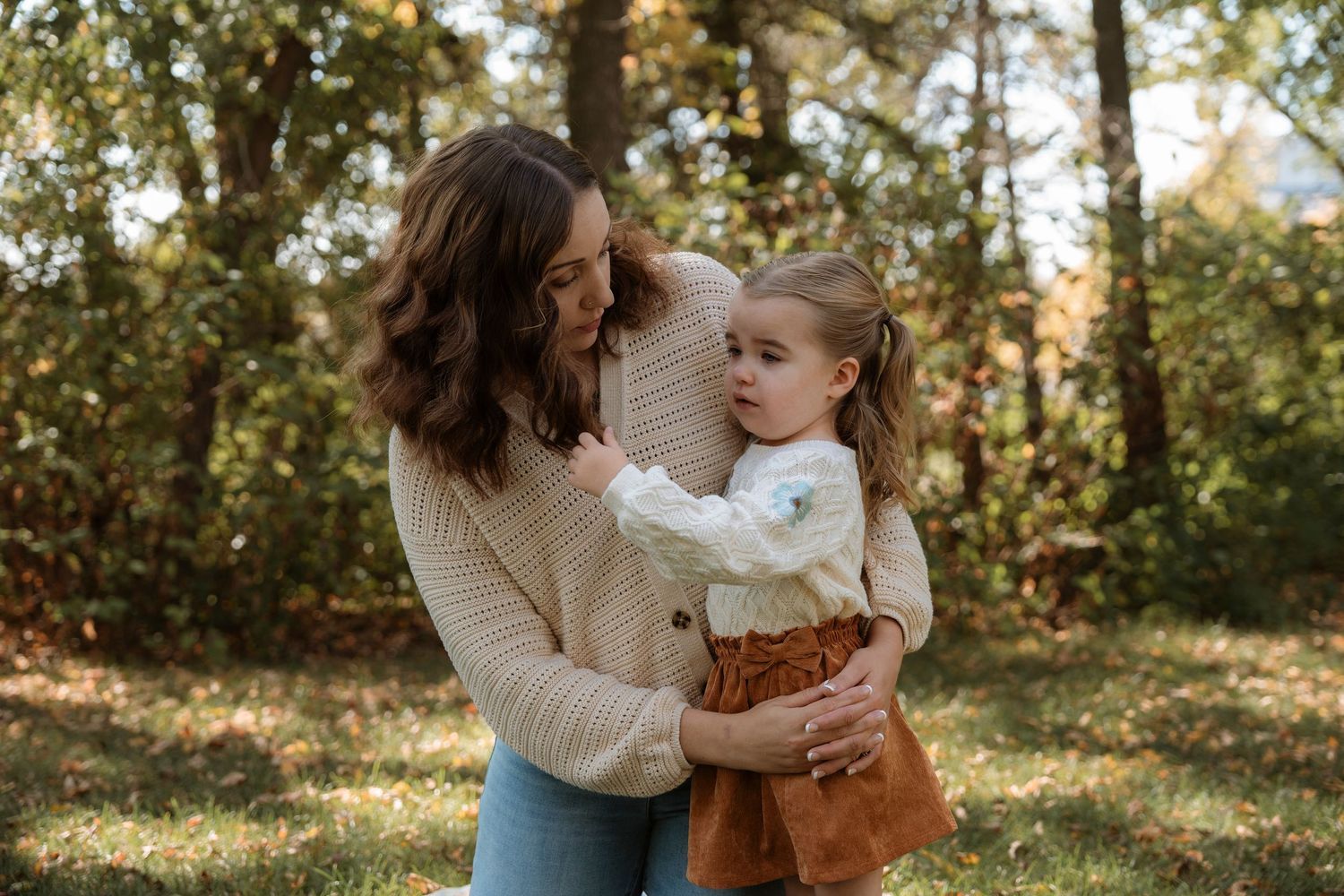 A mother holds her young daughter in an autumn park, both wearing cozy neutral outfits among fall foliage.