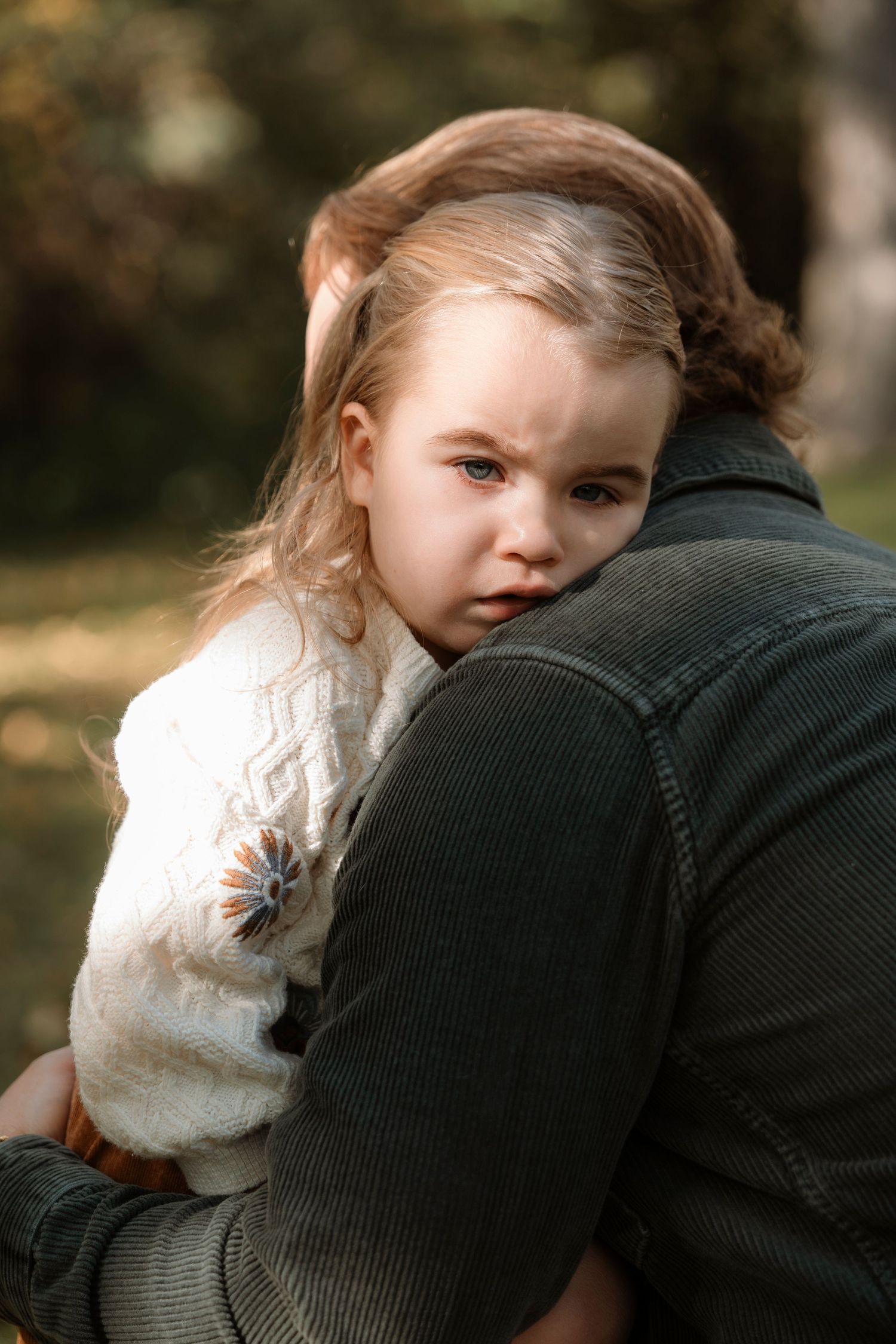 A young girl in a white sweater rests her head on an adult's shoulder outdoors, looking contemplative.