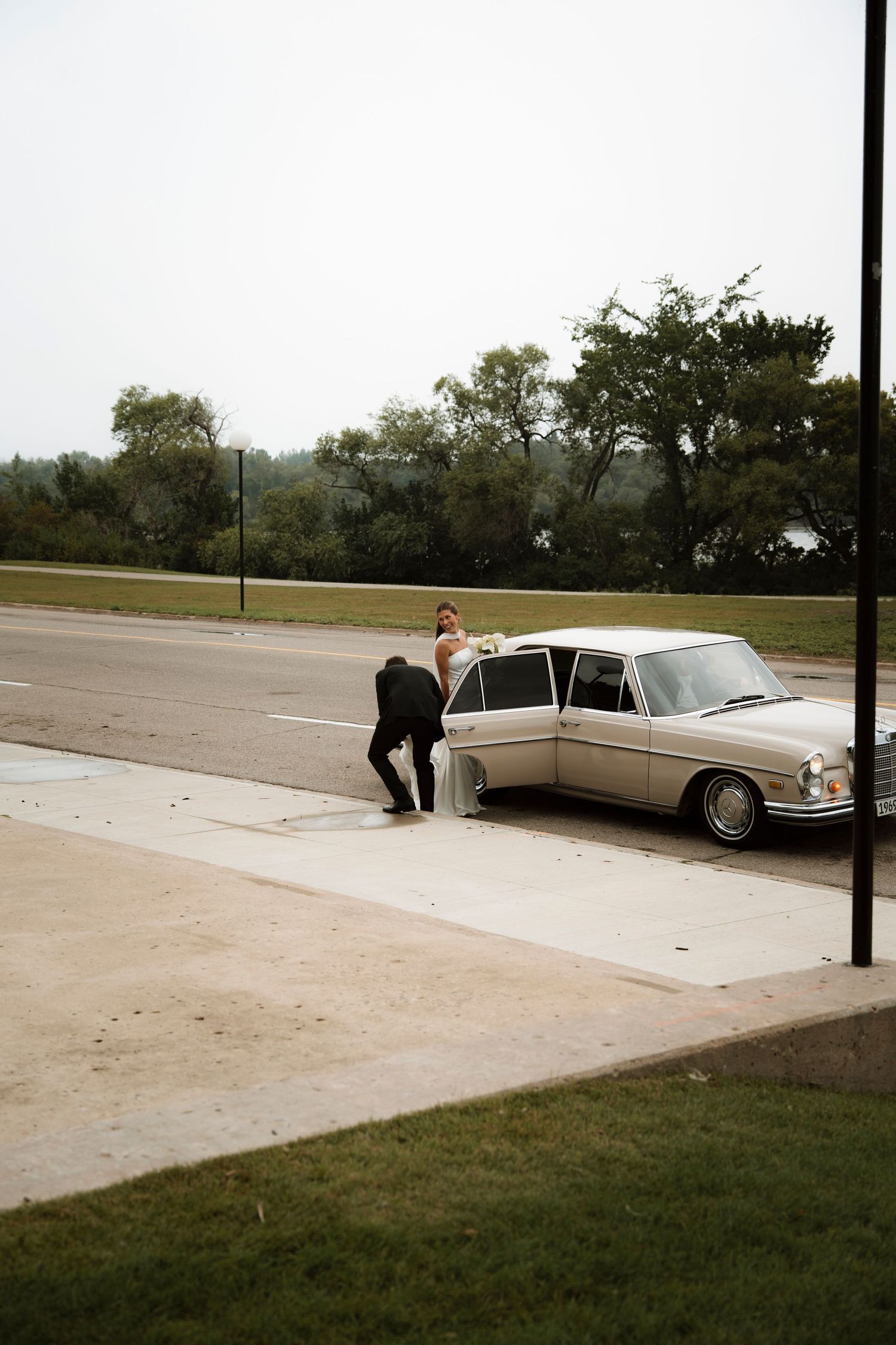 Bride and groom in formal attire beside a vintage cream Mercedes-Benz sedan on a quiet road surrounded by trees.