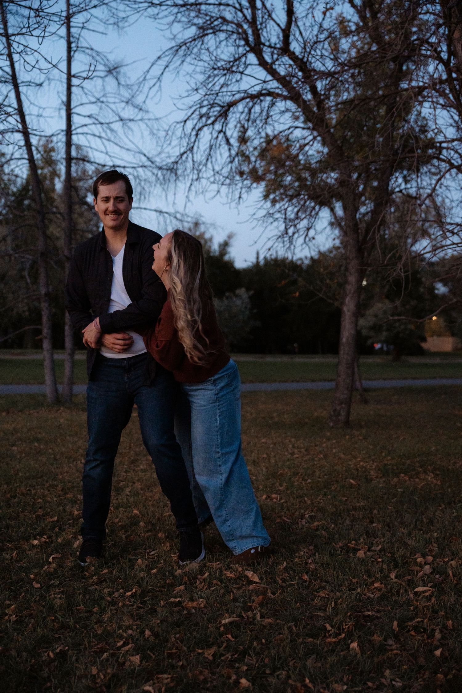 Couple dancing and laughing together in a park at dusk, surrounded by bare trees and fallen autumn leaves.