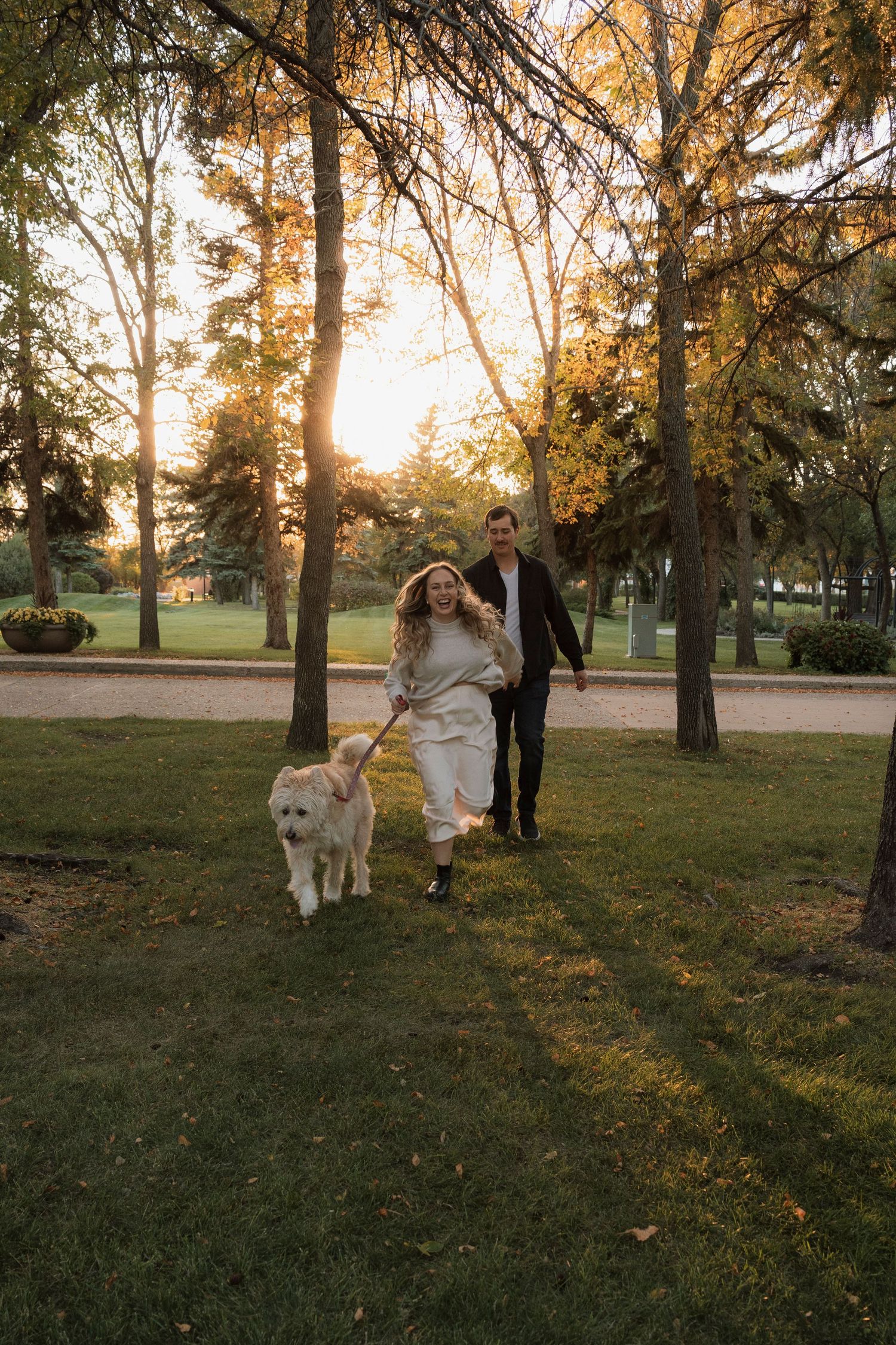 Couple running with a fluffy white dog in a sunlit park during golden hour, surrounded by tall trees.