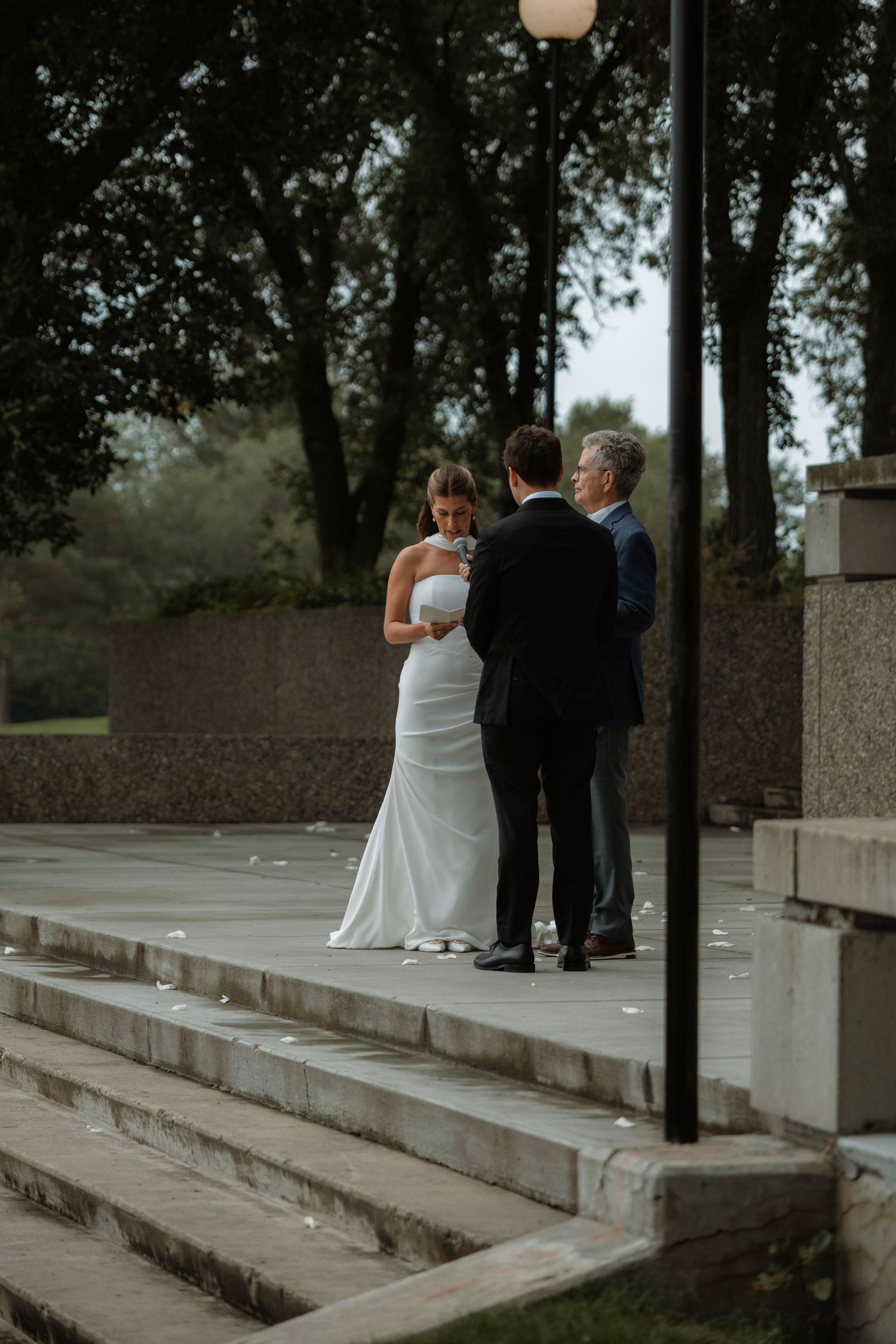 Bride in white gown and groom in black suit exchange vows with officiant on stone steps outdoors.