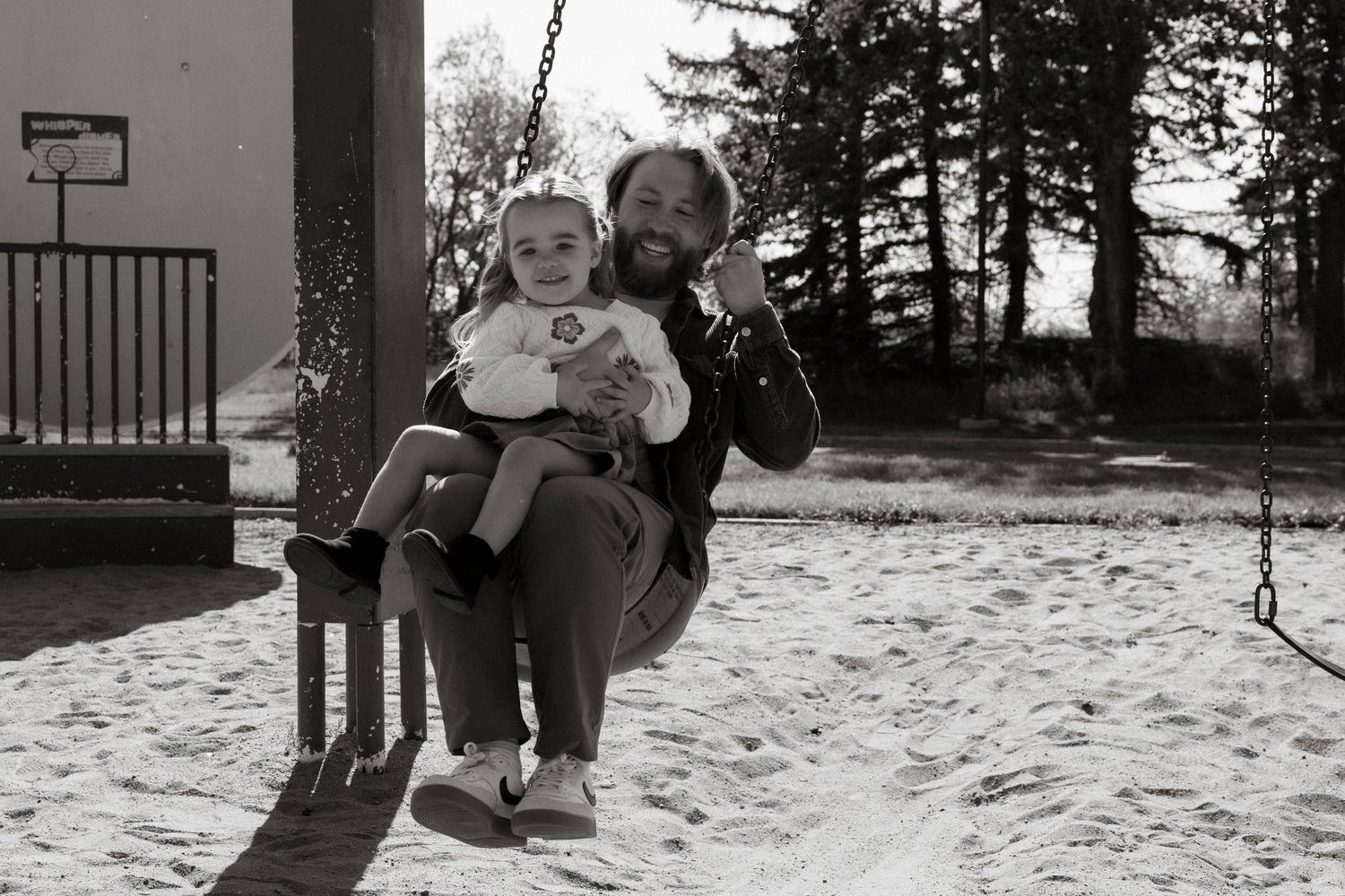 A bearded man sits on a playground swing holding a smiling baby in a sandy play area with trees in the background.