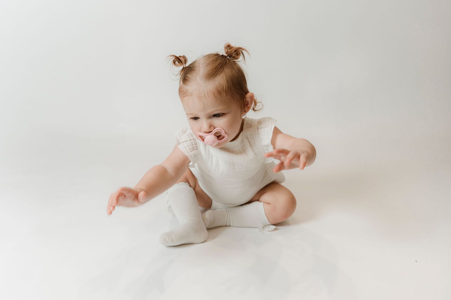 Adorable baby girl in white romper sitting on floor with pigtails, reaching forward with a playful expression.