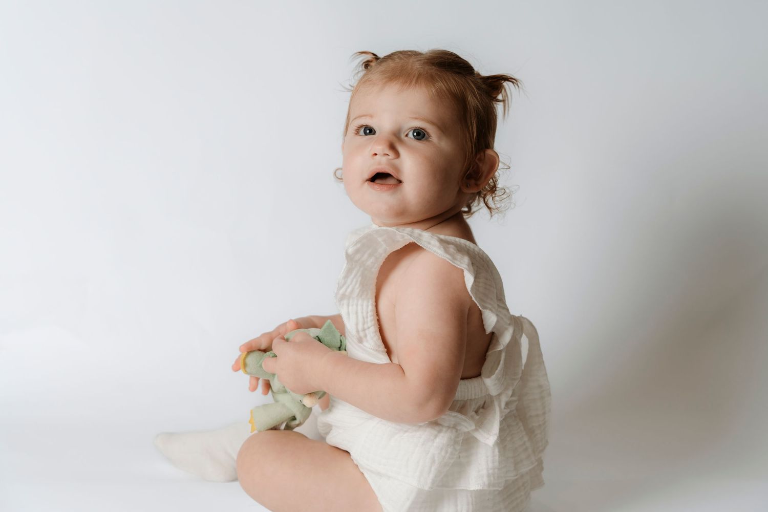 Adorable baby girl in white lace romper sitting on white background, looking up curiously while holding a toy.