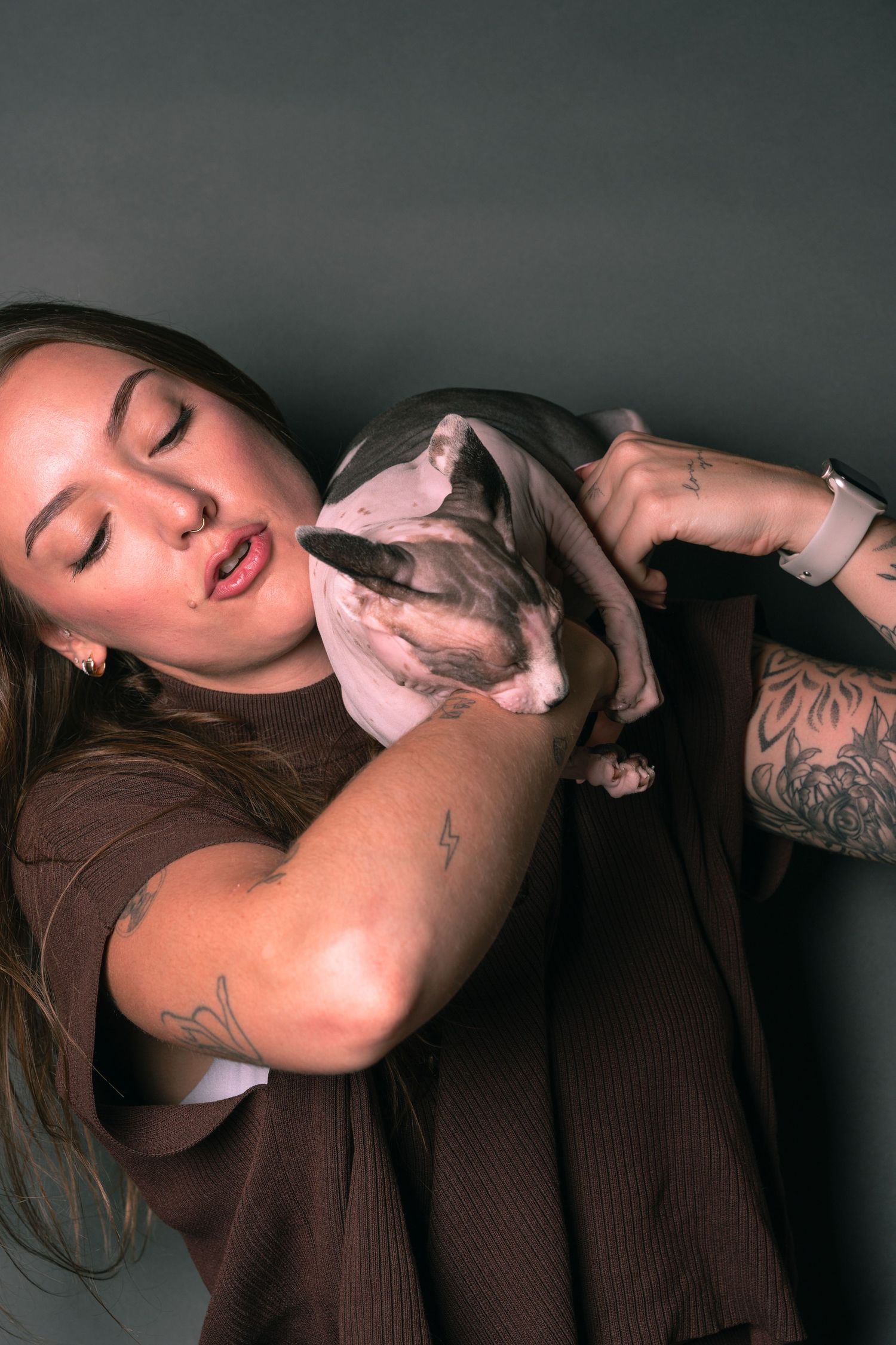 Tattooed woman with long hair holding a Sphynx cat close to her chest against a dark gray background.