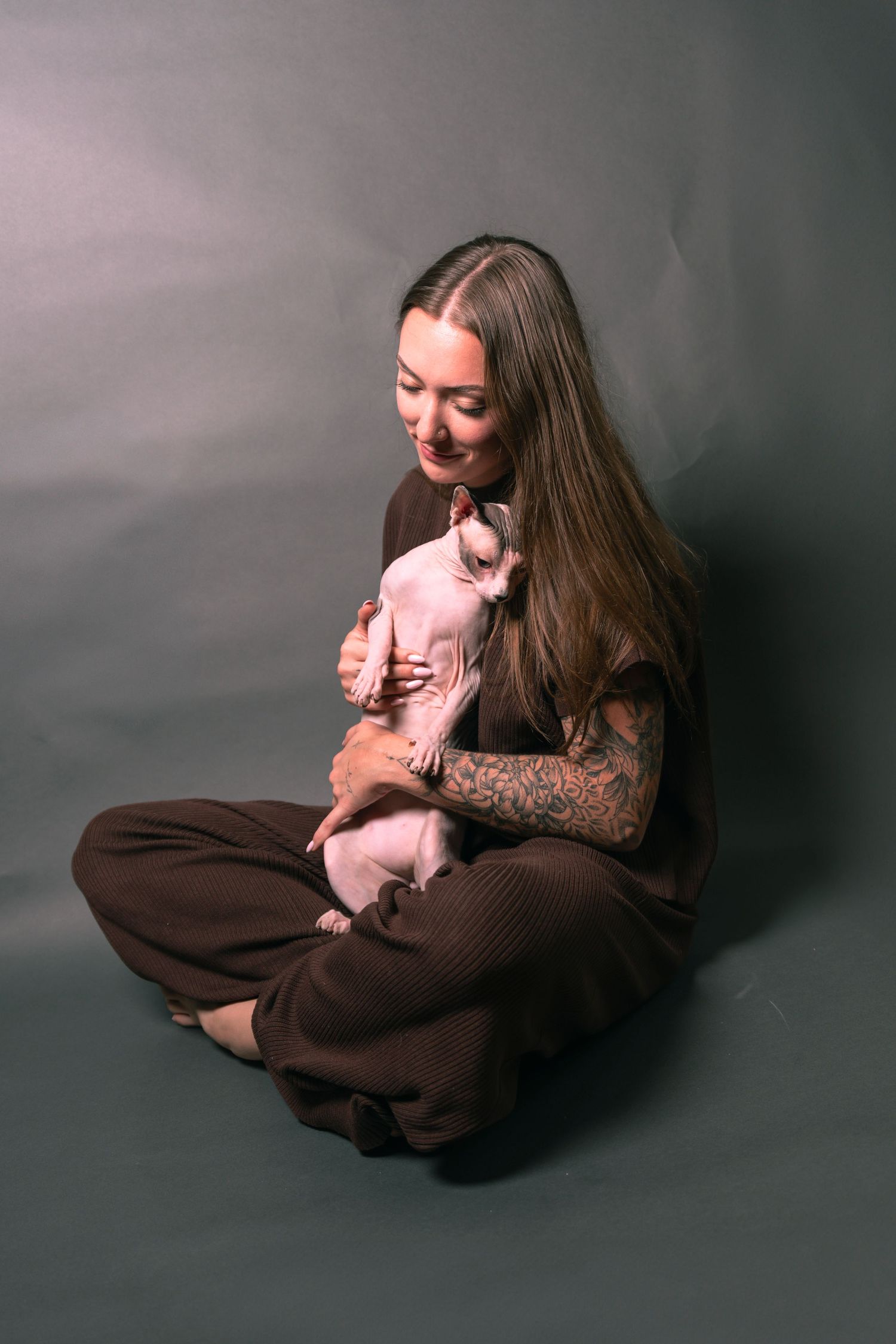 Woman with tattoos sitting cross-legged on gray background, gently holding a hairless Sphynx cat in her arms.