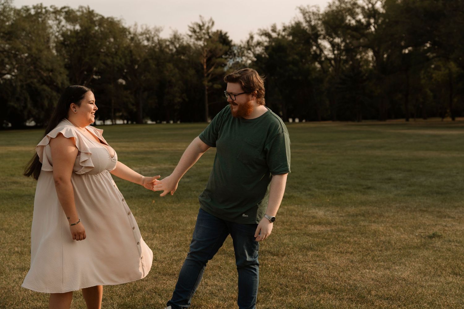 Couple holding hands and dancing outdoors on green grass field during golden hour engagement photo session.
