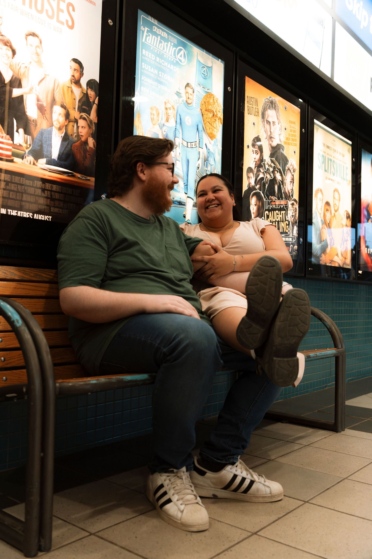 Couple laughing together on a bench outside a movie theater with film posters displayed on the wall behind them.