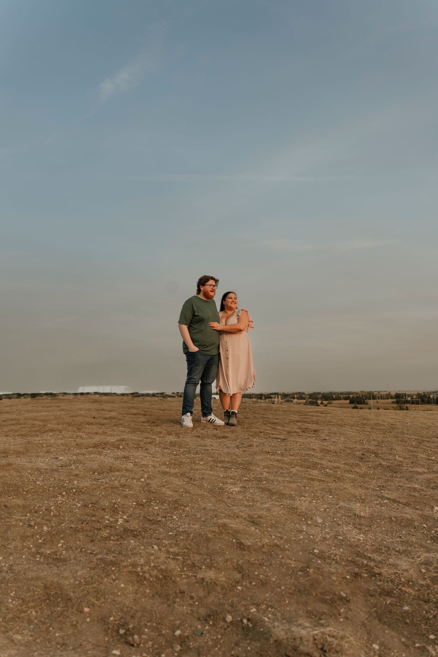 A couple stands embracing on an open dirt field under a vast sky at golden hour, wearing casual attire.