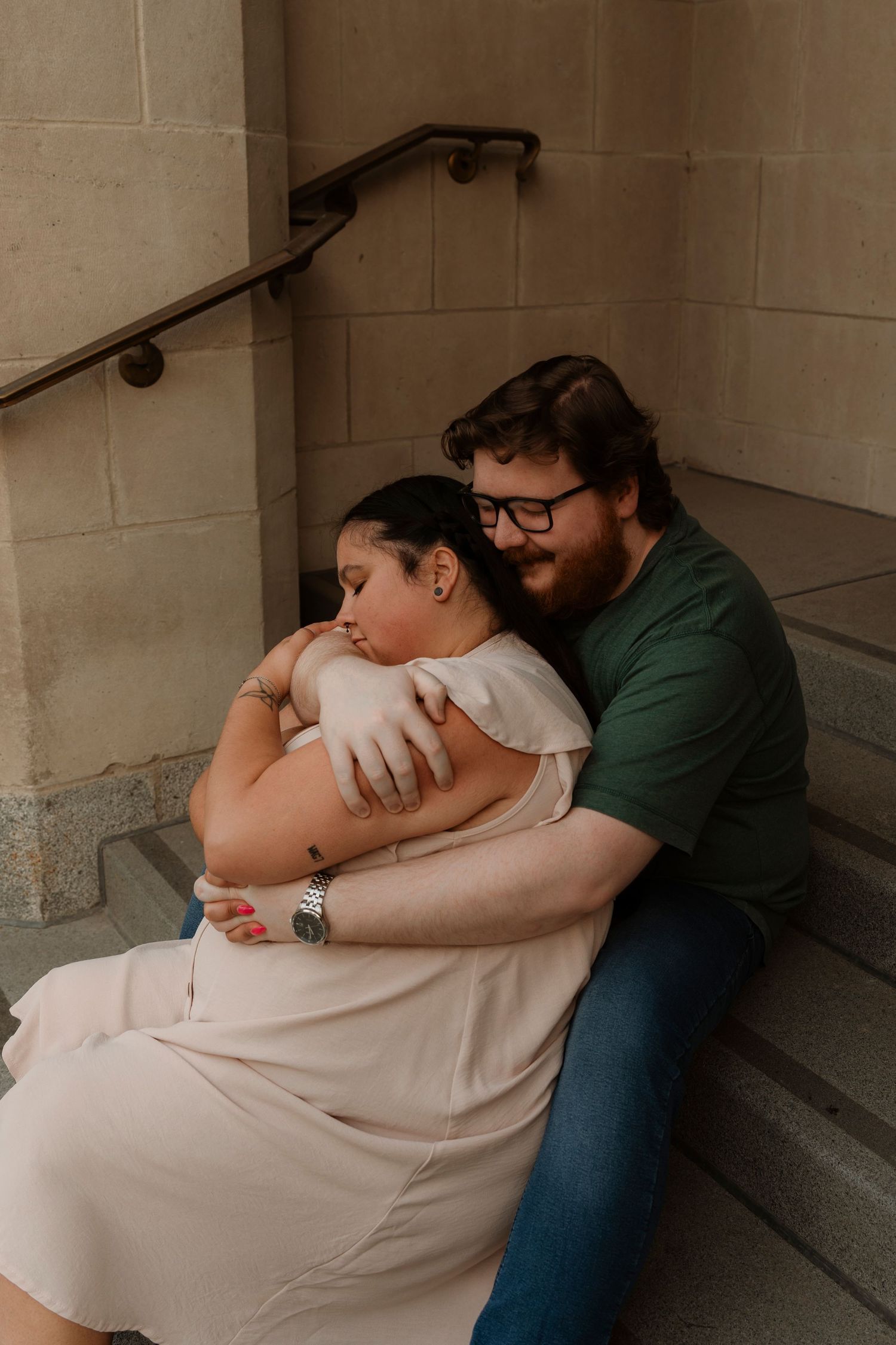 A couple sits on stone steps, the man in a green shirt embracing a woman in a blush dress, sharing a tender moment.