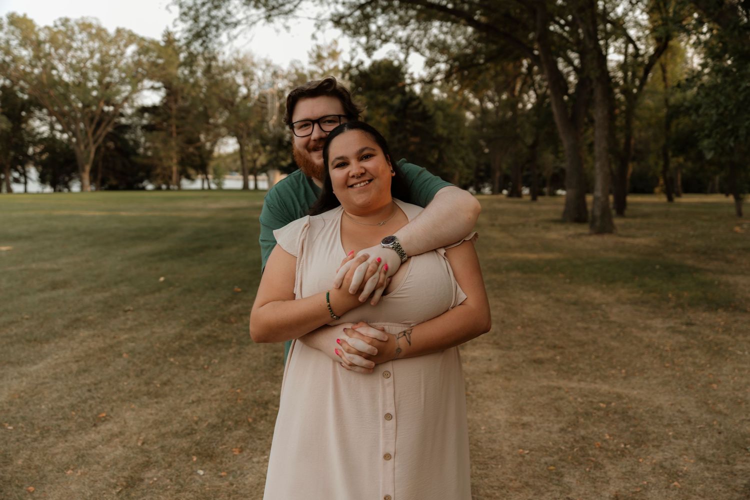 Couple embracing and smiling in a lush green park during golden hour engagement photo session.