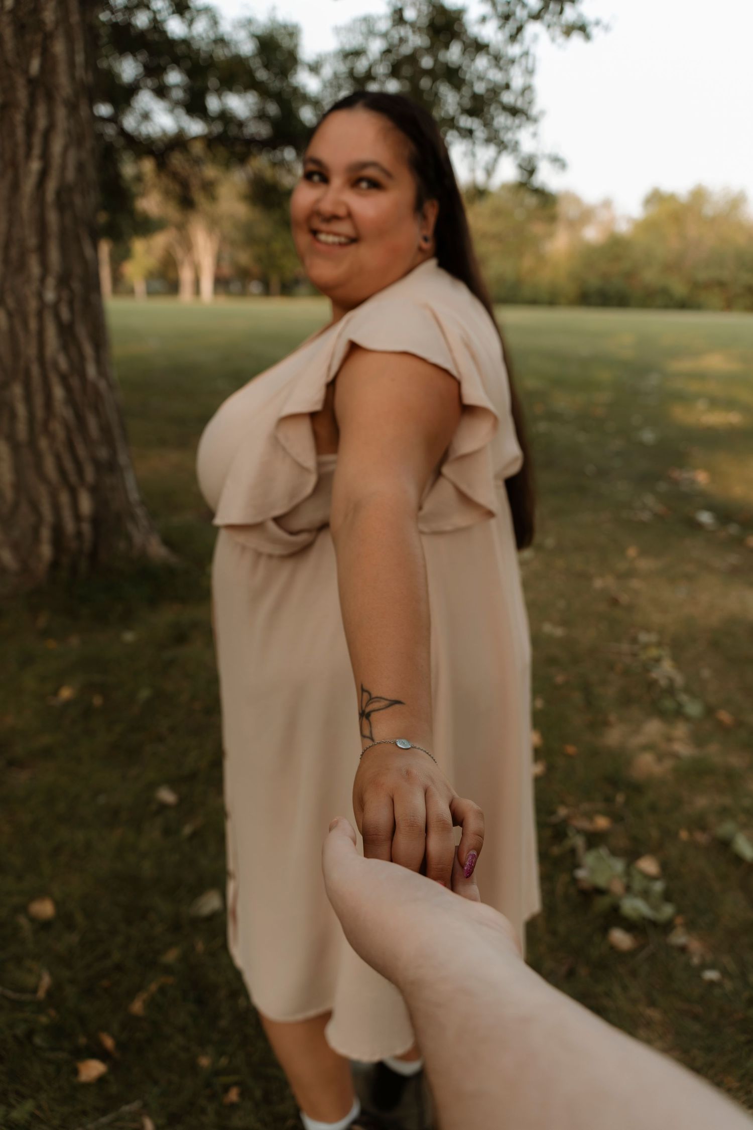 Smiling woman in beige ruffle dress holds hands with partner in a sunny park setting.