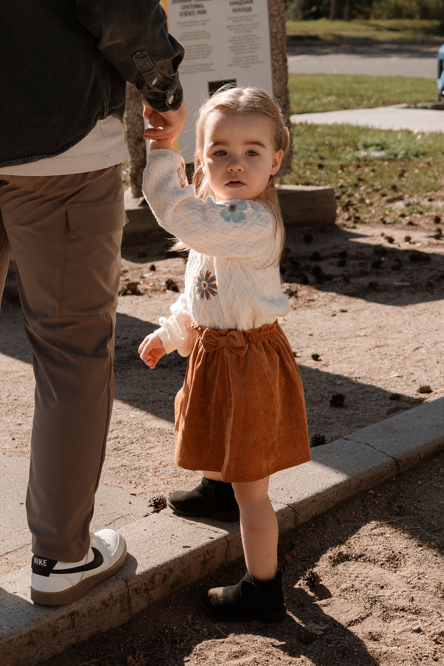 Toddler girl in white sweater and brown skirt holds adult's hand outdoors on a sunny day.
